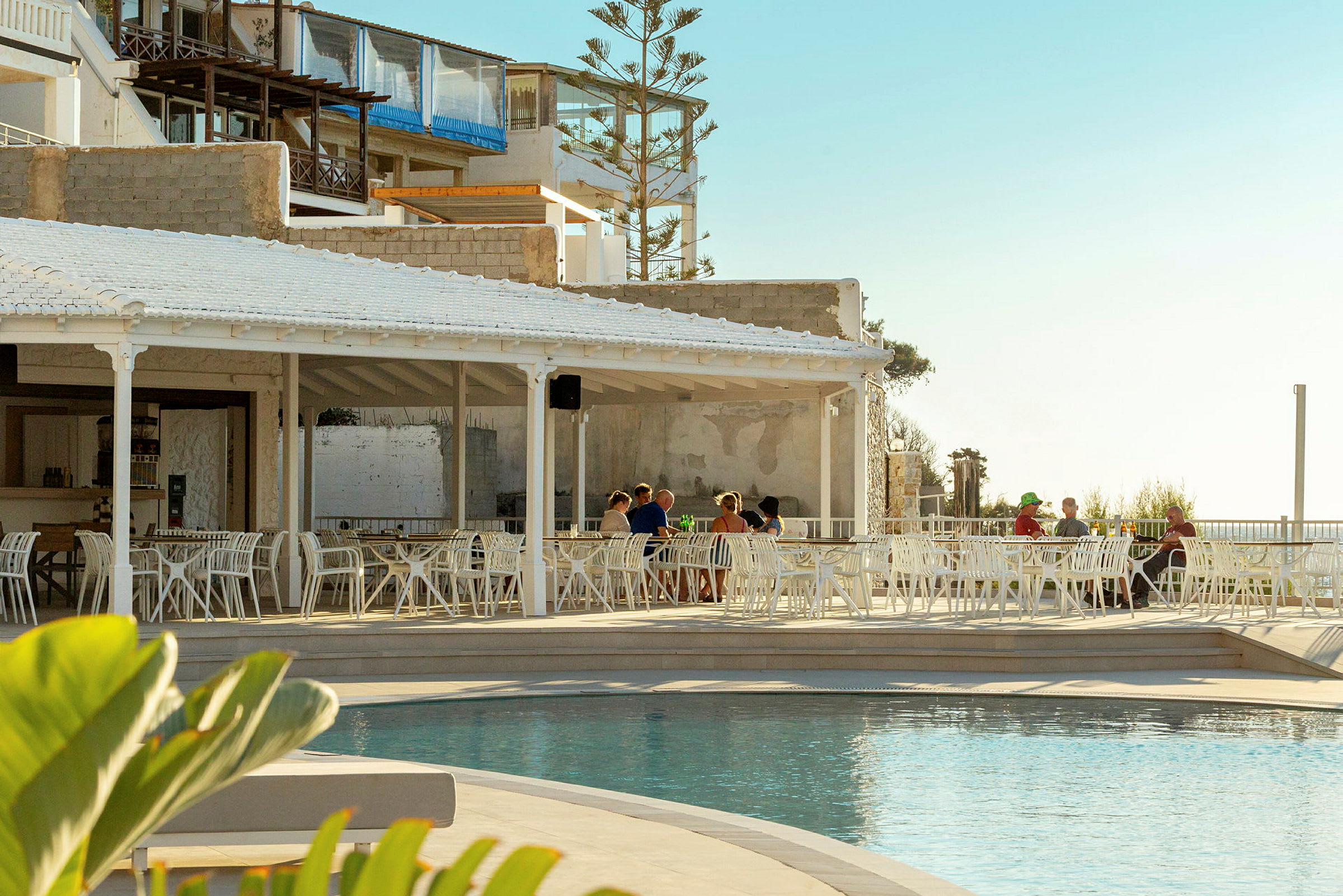 En udendørs restaurant med hvide møbler ved siden af en pool, med nogle personer der sidder ved bordene. Baggrunden består af bygninger og en lys himmel. (AI-genereret tekst)