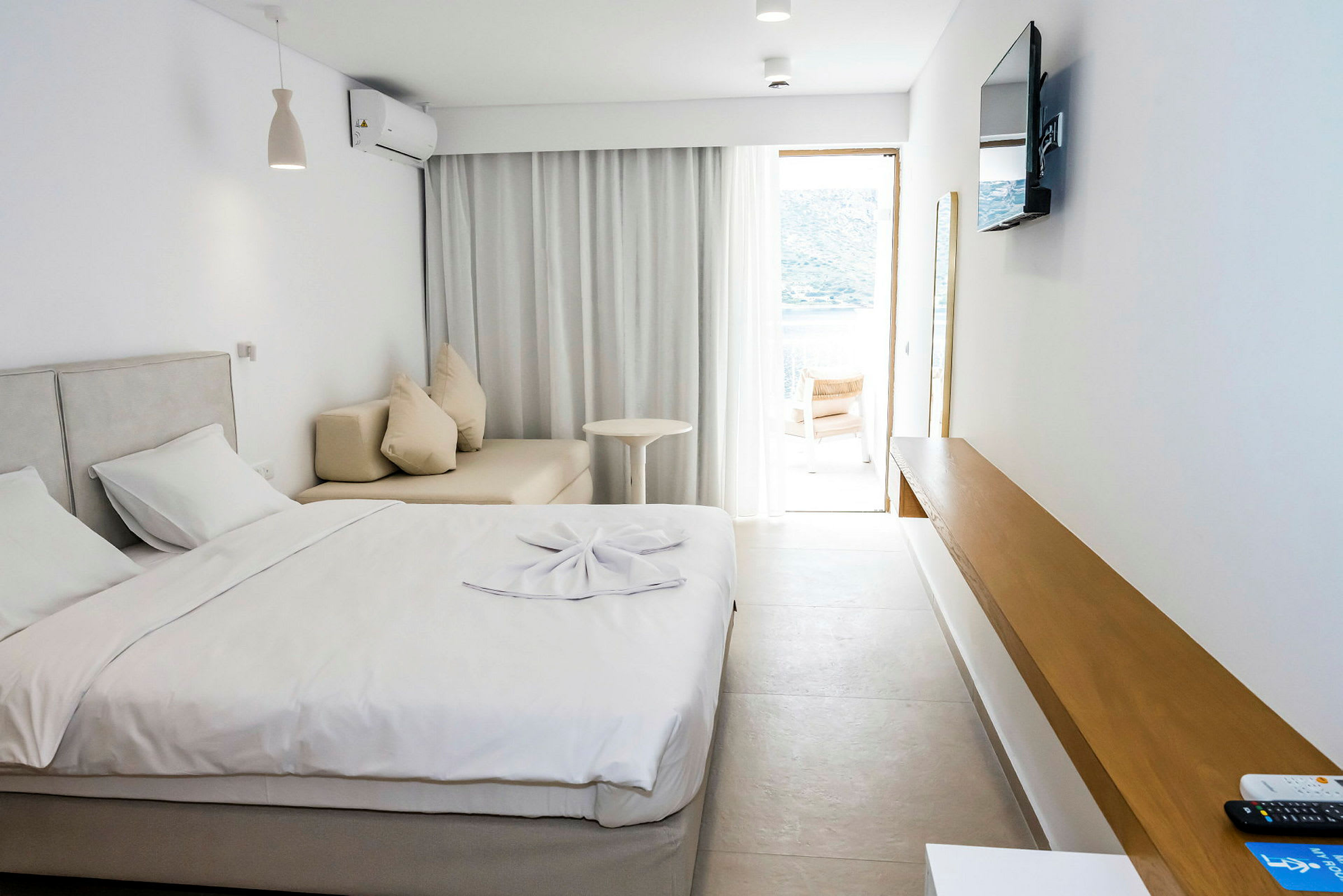 Et hotelværelse med en seng, sofa, et bord og et tv på væggen. Et vindue med lyse gardiner fører ud til en terrasse. (AI-genereret tekst)