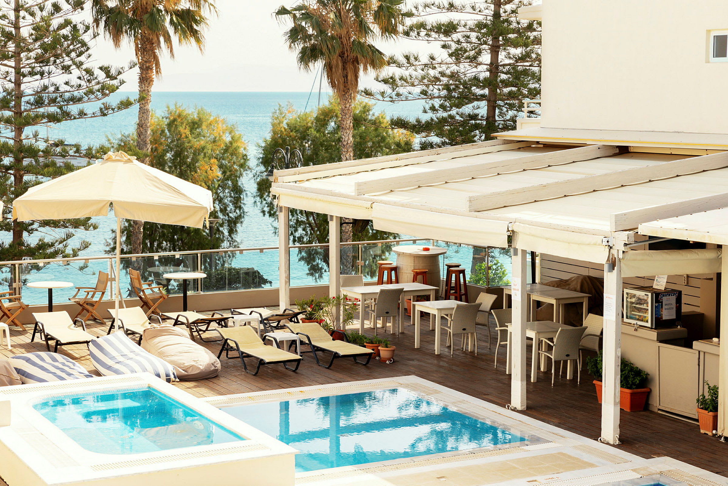 Billede af en udendørs pool med liggestole og parasoller, samt en terrasse med møbler og udsigt over havet. (AI-genereret tekst)