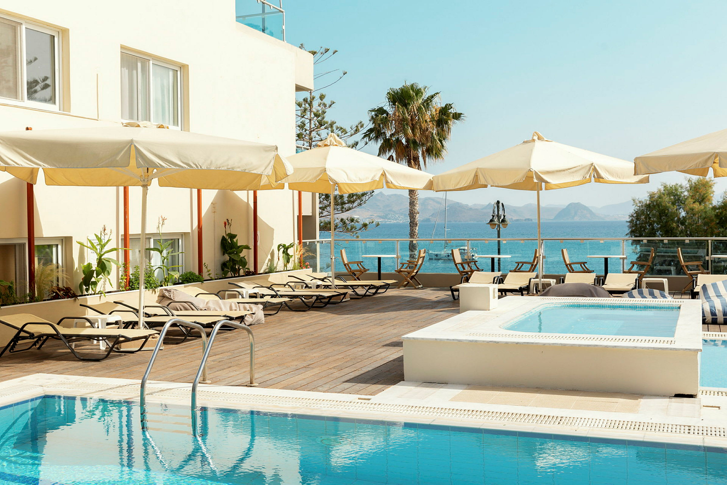 Poolområde ved hotel med liggestole og parasoller, havet og bjerge i baggrunden. (AI-genereret tekst)