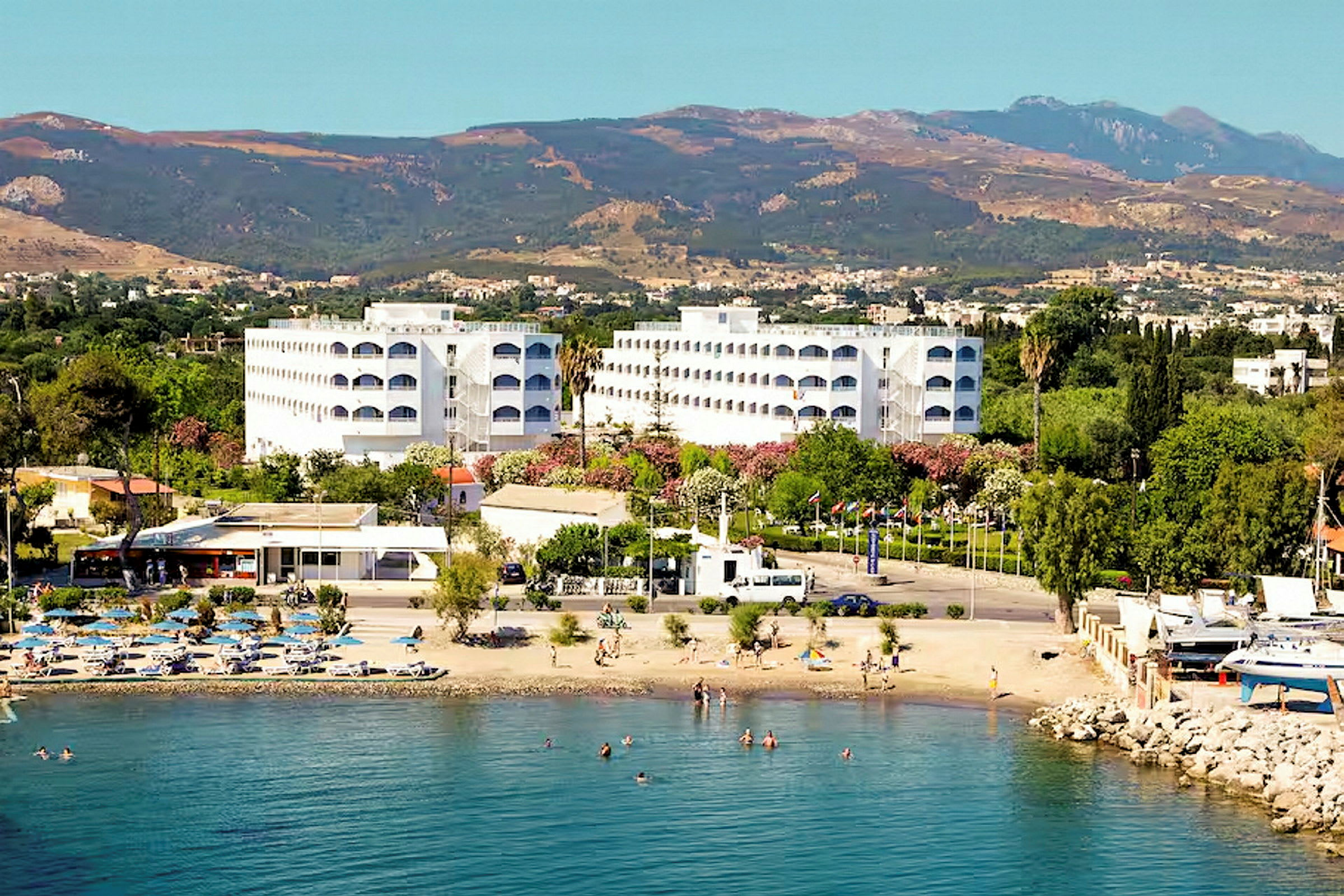 Hotelbygninger ved en strand med badende personer og bjerge i baggrunden. (AI-genereret tekst)