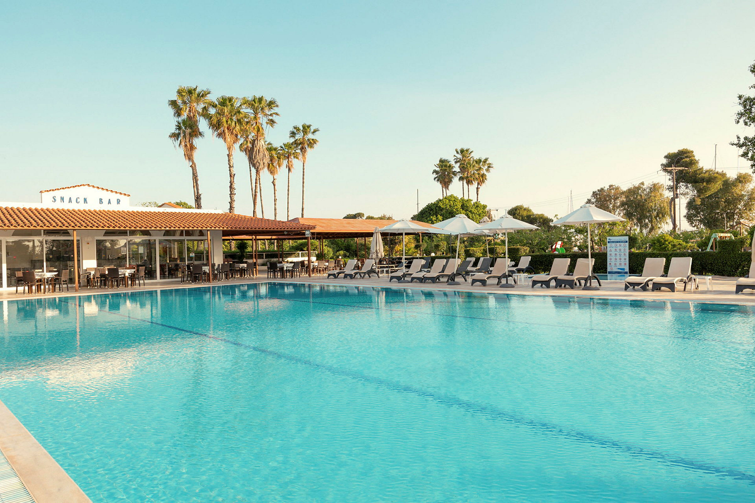 Udendørs swimmingpool med liggestole og parasoller på siden. Bagved ses en bygning med skiltet 'SNACK BAR' og palmer i baggrunden. (AI-genereret tekst)