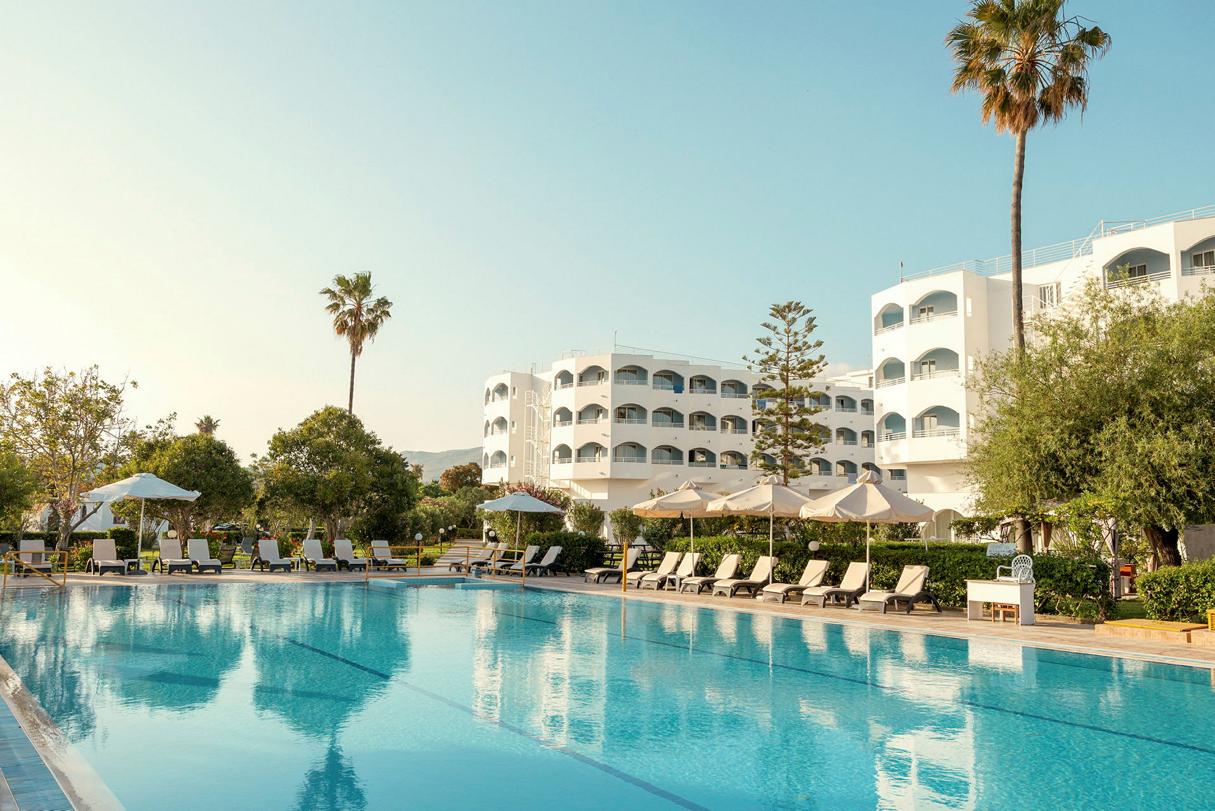 Hotelbygning med pool, liggestole og parasoller i forgrunden. (AI-genereret tekst)