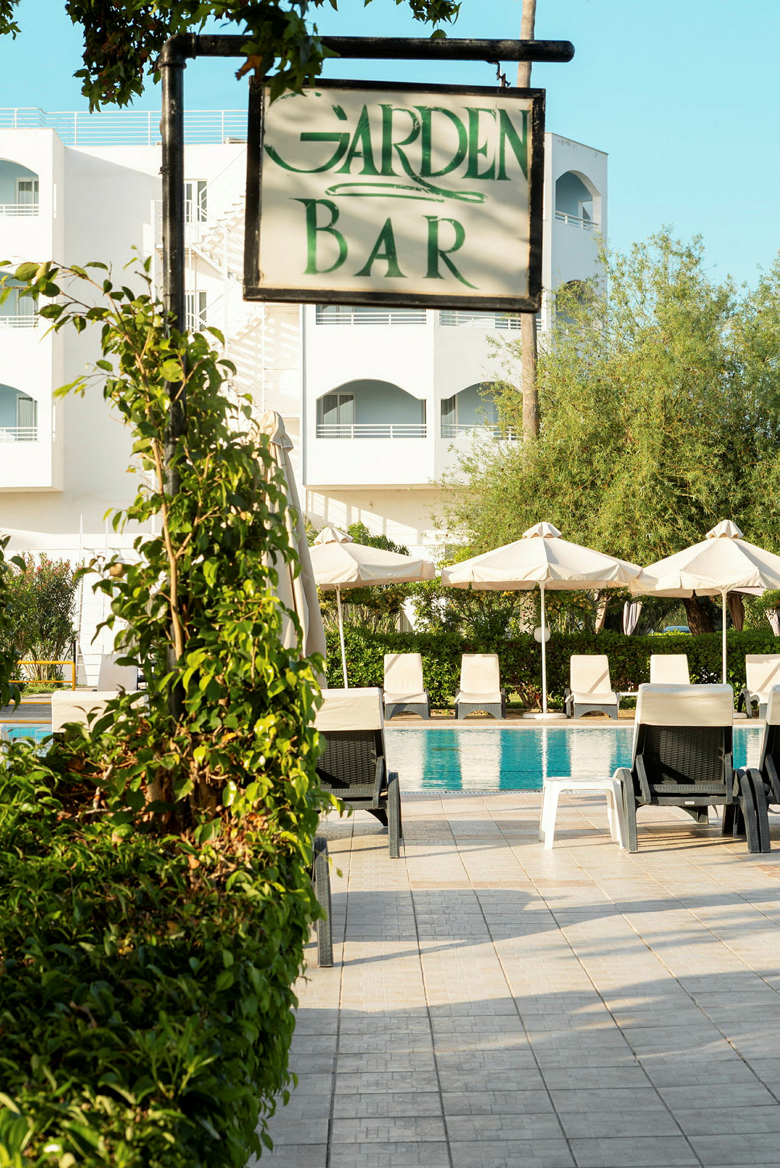 Skilt med teksten 'Garden Bar' foran poolområde og hotelbygning. (AI-genereret tekst)