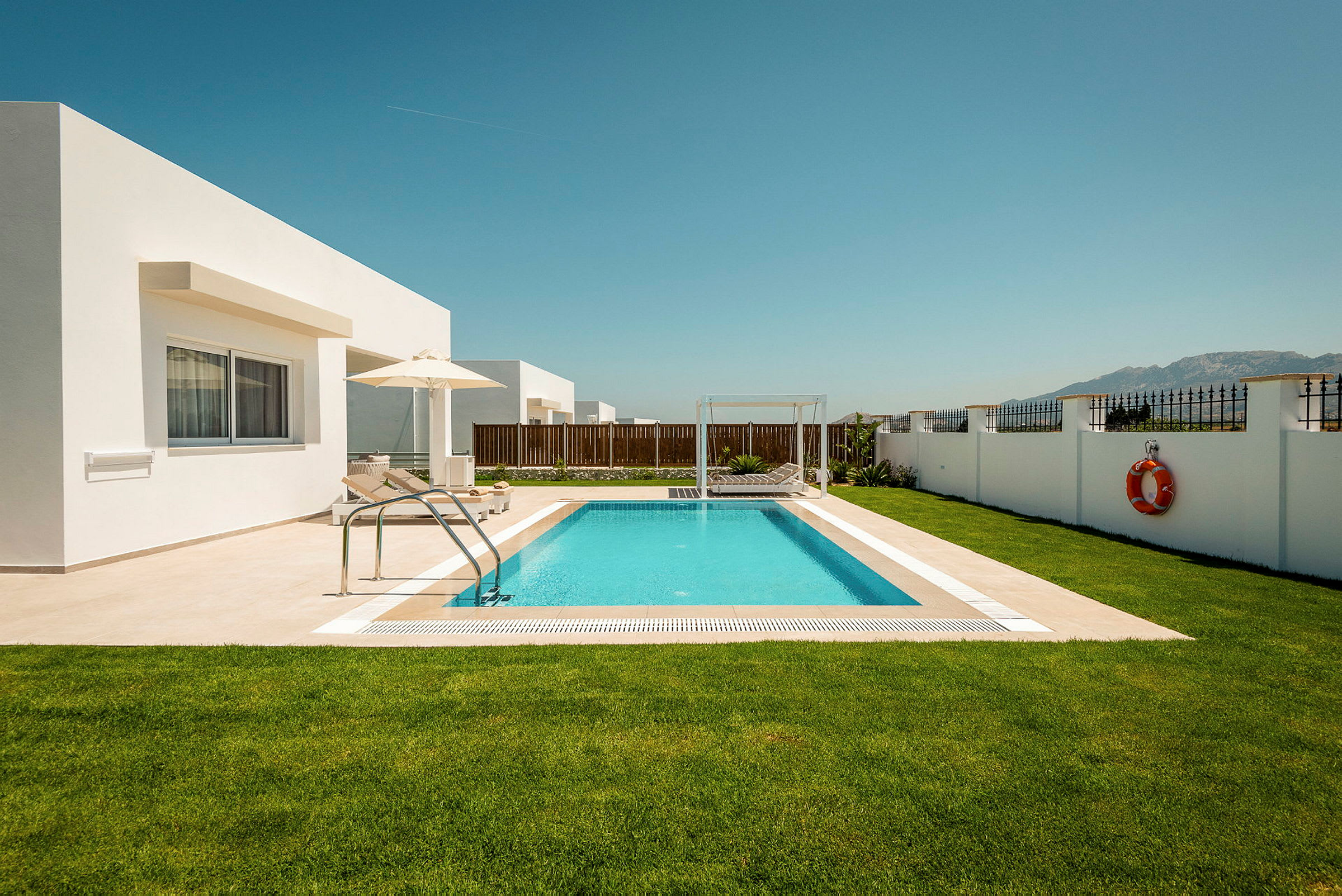 En privat pool med liggestole og parasol omgivet af en græsplæne og en hvid mur. Moderne arkitektur og en klar blå himmel. (AI-genereret tekst)