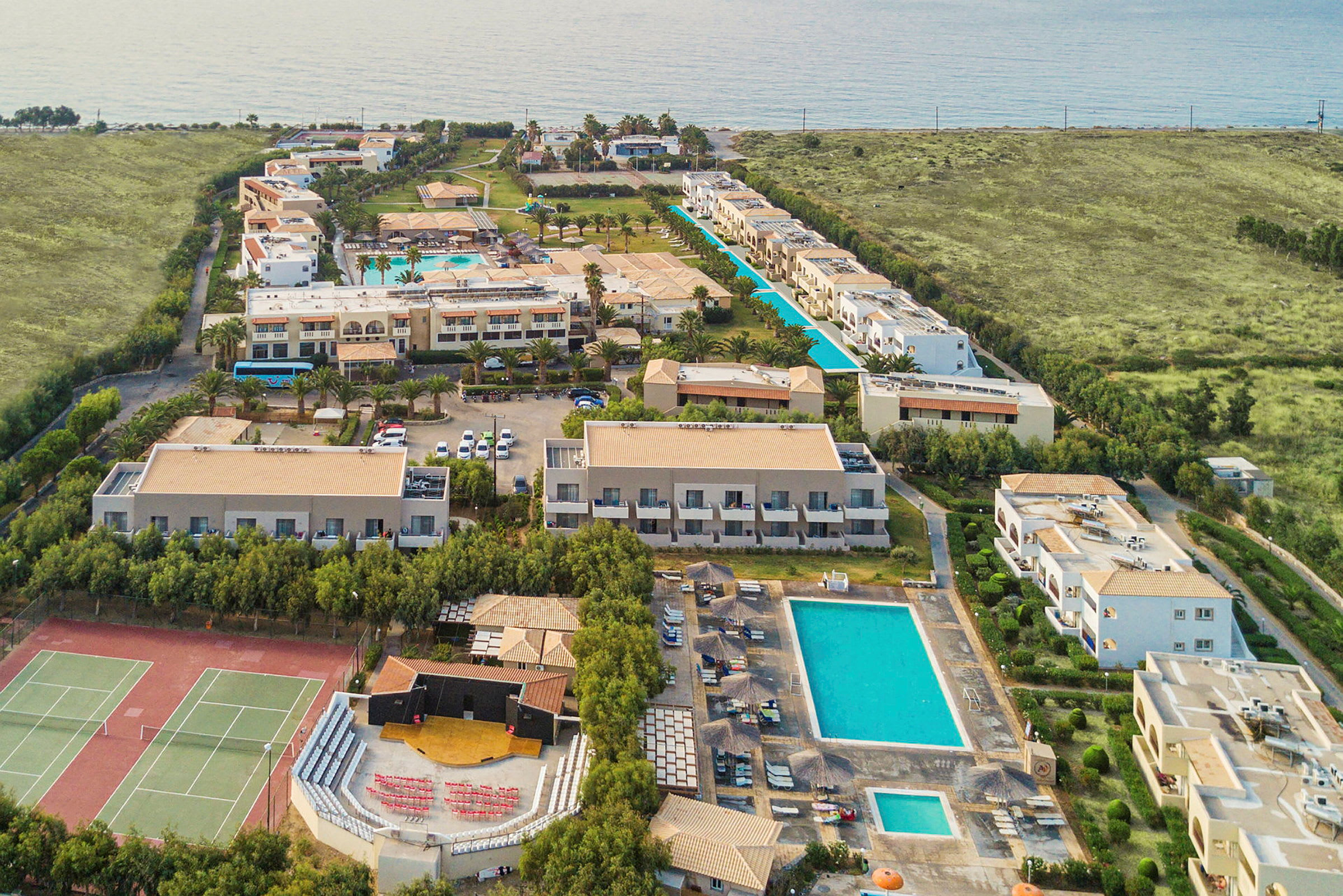 Luftfoto af et feriested med bygninger, swimmingpools og tennisbaner nær kysten. (AI-genereret tekst)