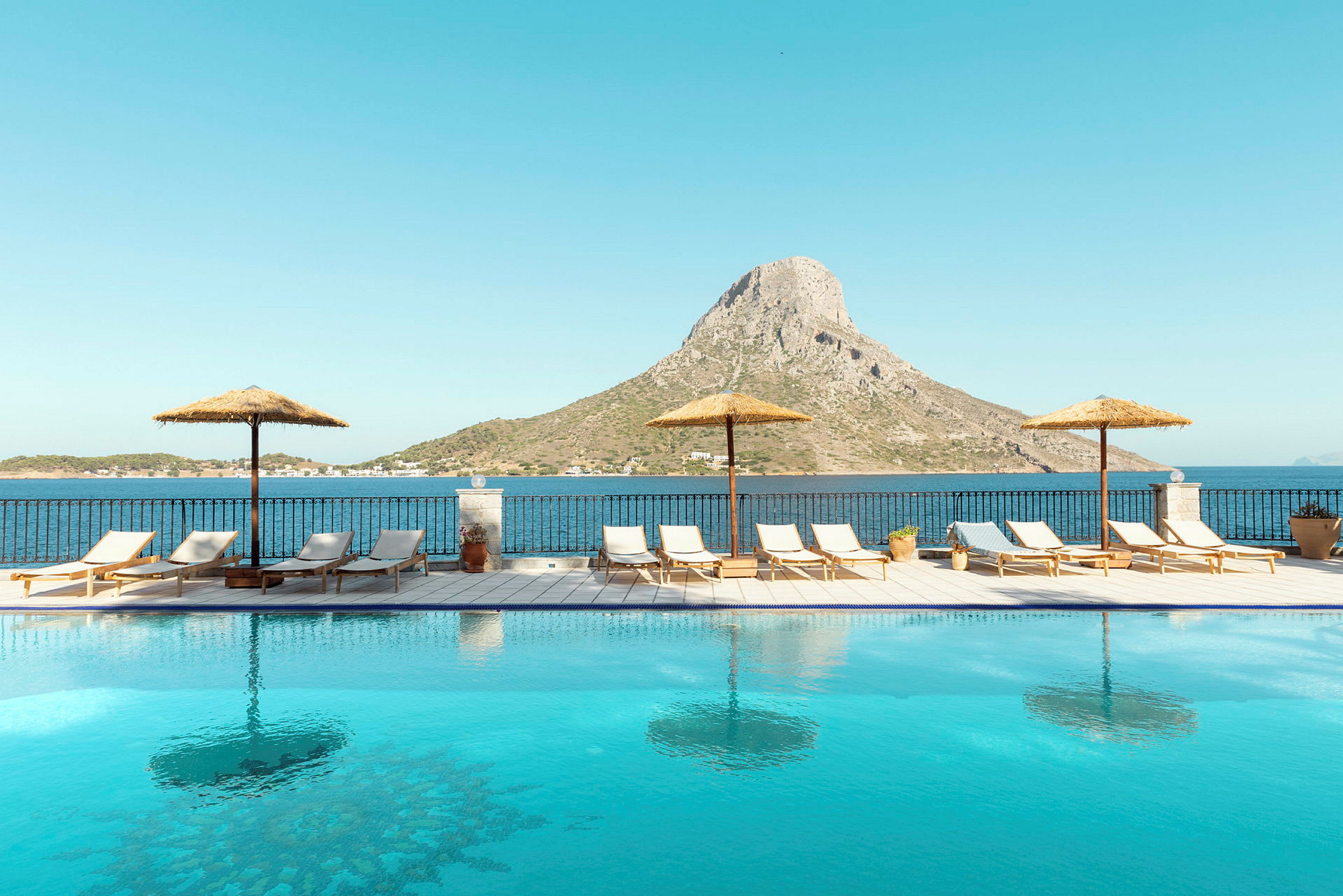 Udendørs pool med liggestole og parasoller med udsigt over havet og et bjerg. (AI-genereret tekst)
