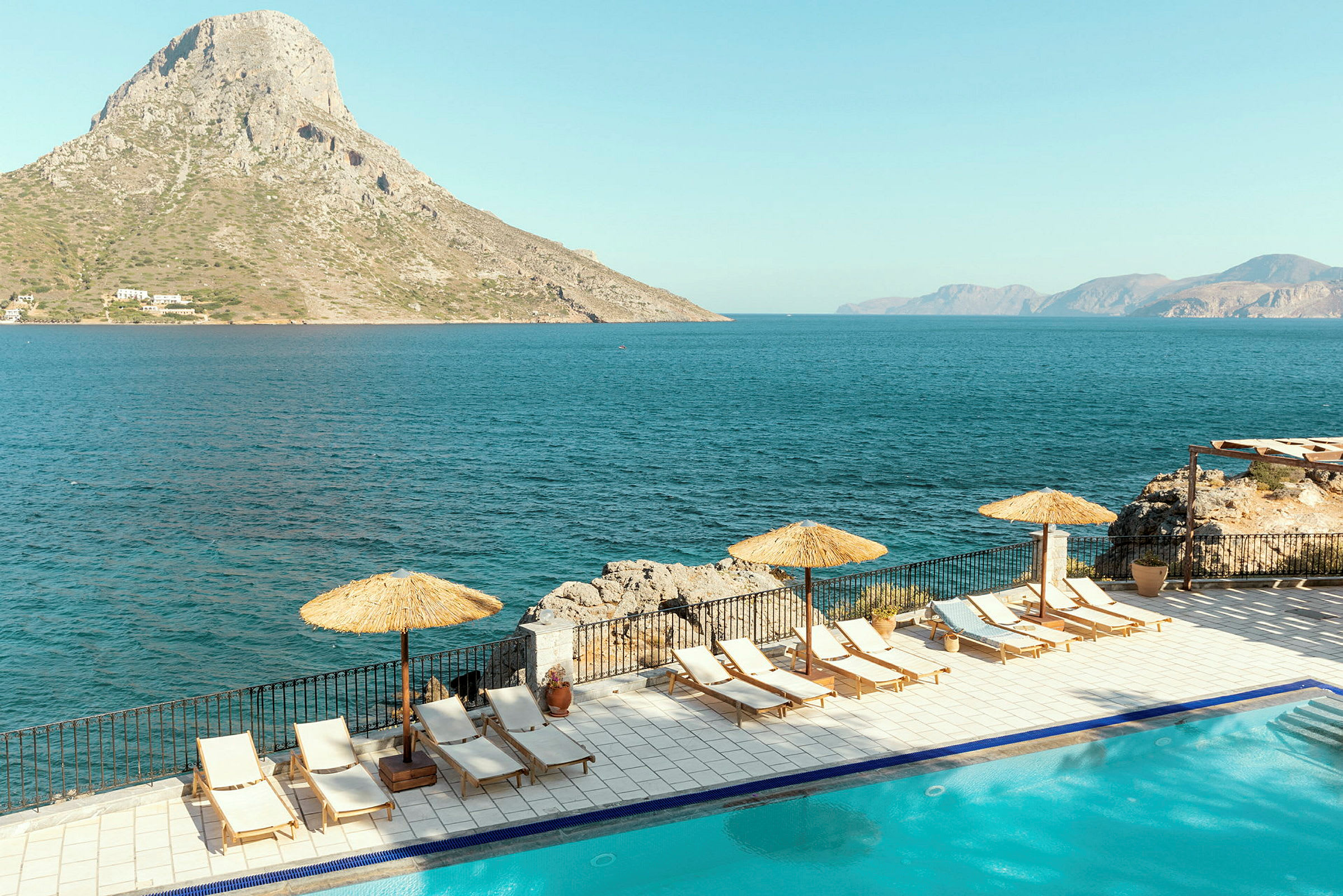 Solstole og parasoller ved en pool med havet i baggrunden. En bakke rejser sig tæt ved stranden. (AI-genereret tekst)