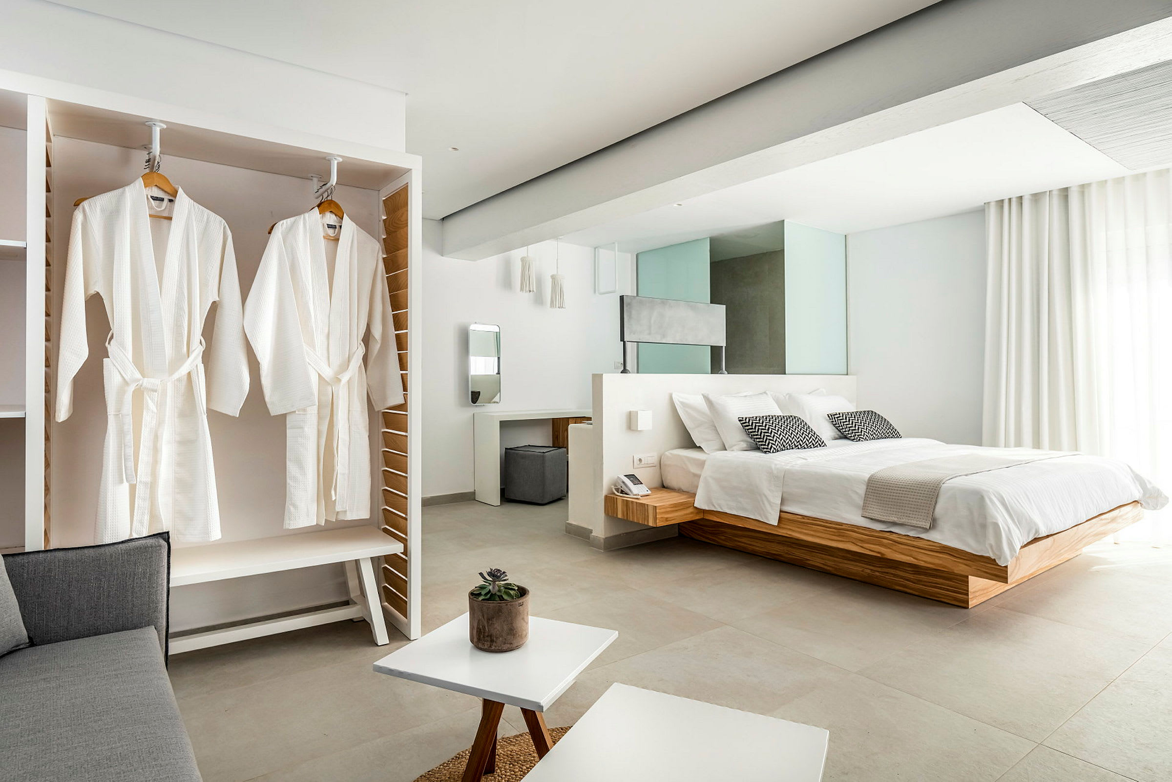 Billede af et elegant hotelværelse med en stor seng, hvide badekåber hængende i et skab og lyse moderne møbler. (AI-genereret tekst)