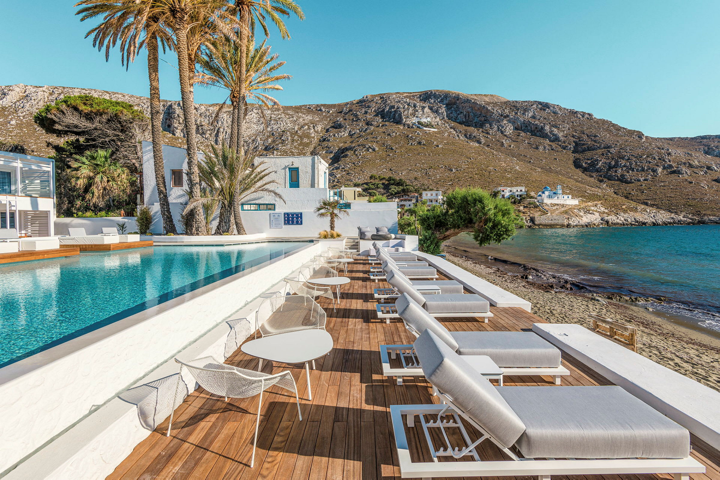 Udendørs swimmingpool med afslapningsstole og udsigt til havet og bjergene i baggrunden. (AI-genereret tekst)
