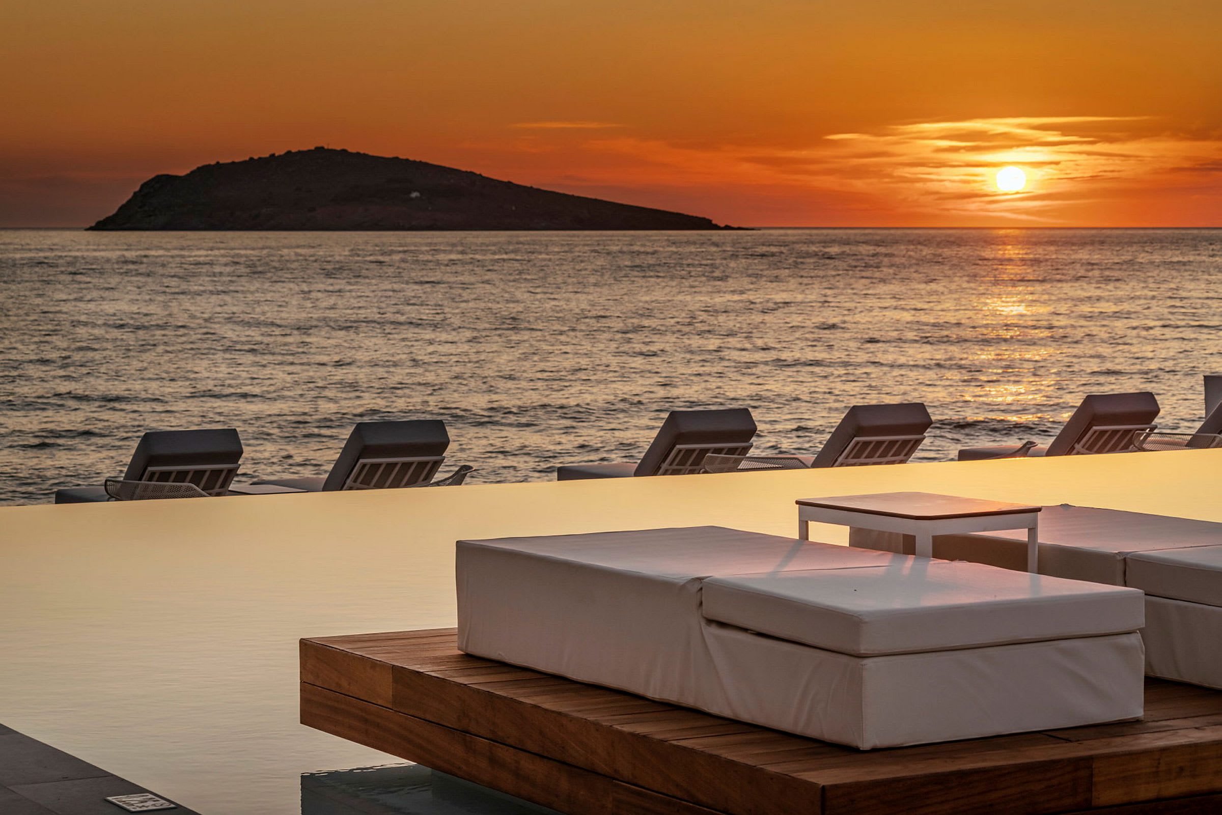 Solnedgang over havet med liggestole ved en infinitypool. (AI-genereret tekst)