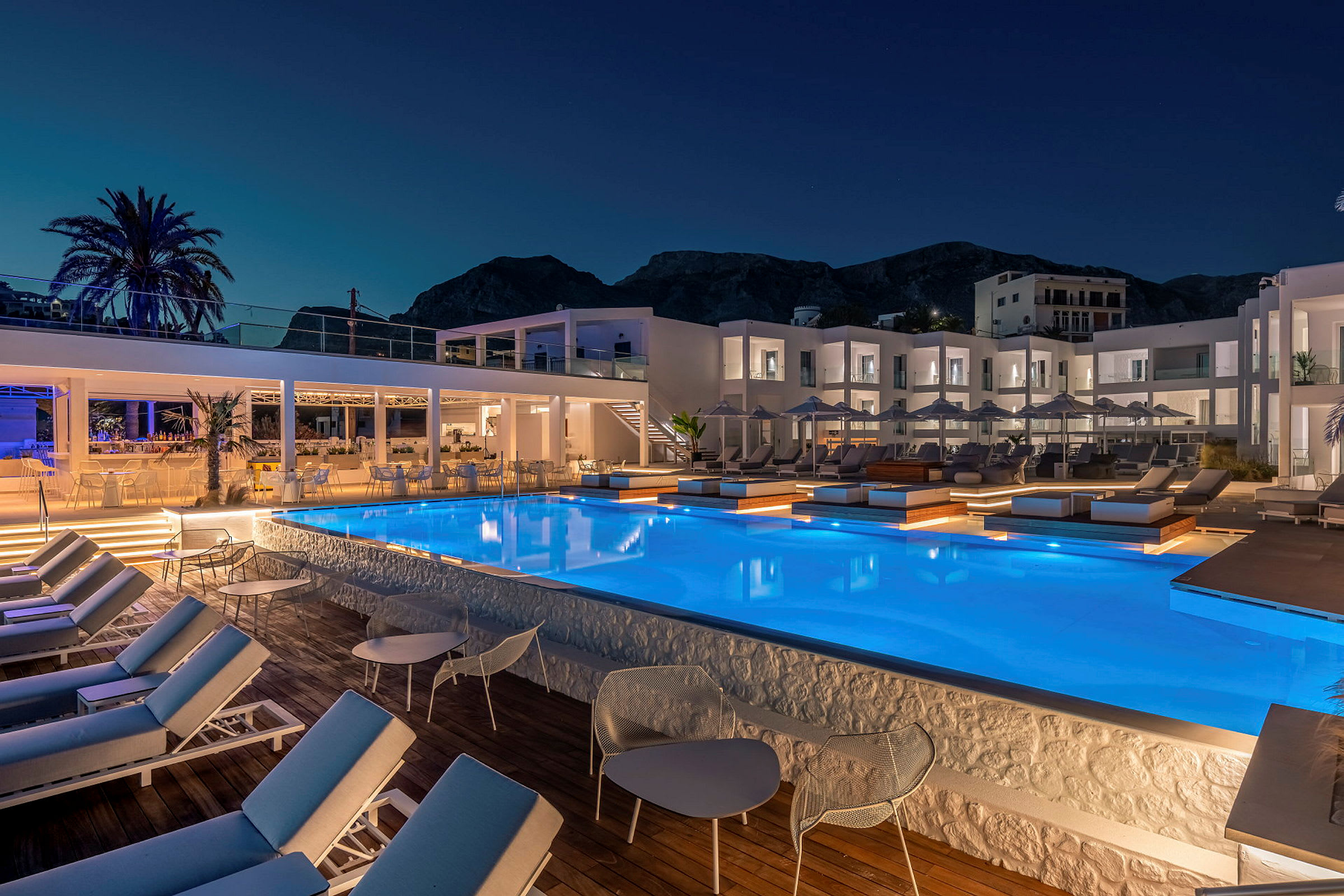 Billede af en oplyst hotelpool med solsenge og omkringliggende bygninger i aftenskumringen. (AI-genereret tekst)