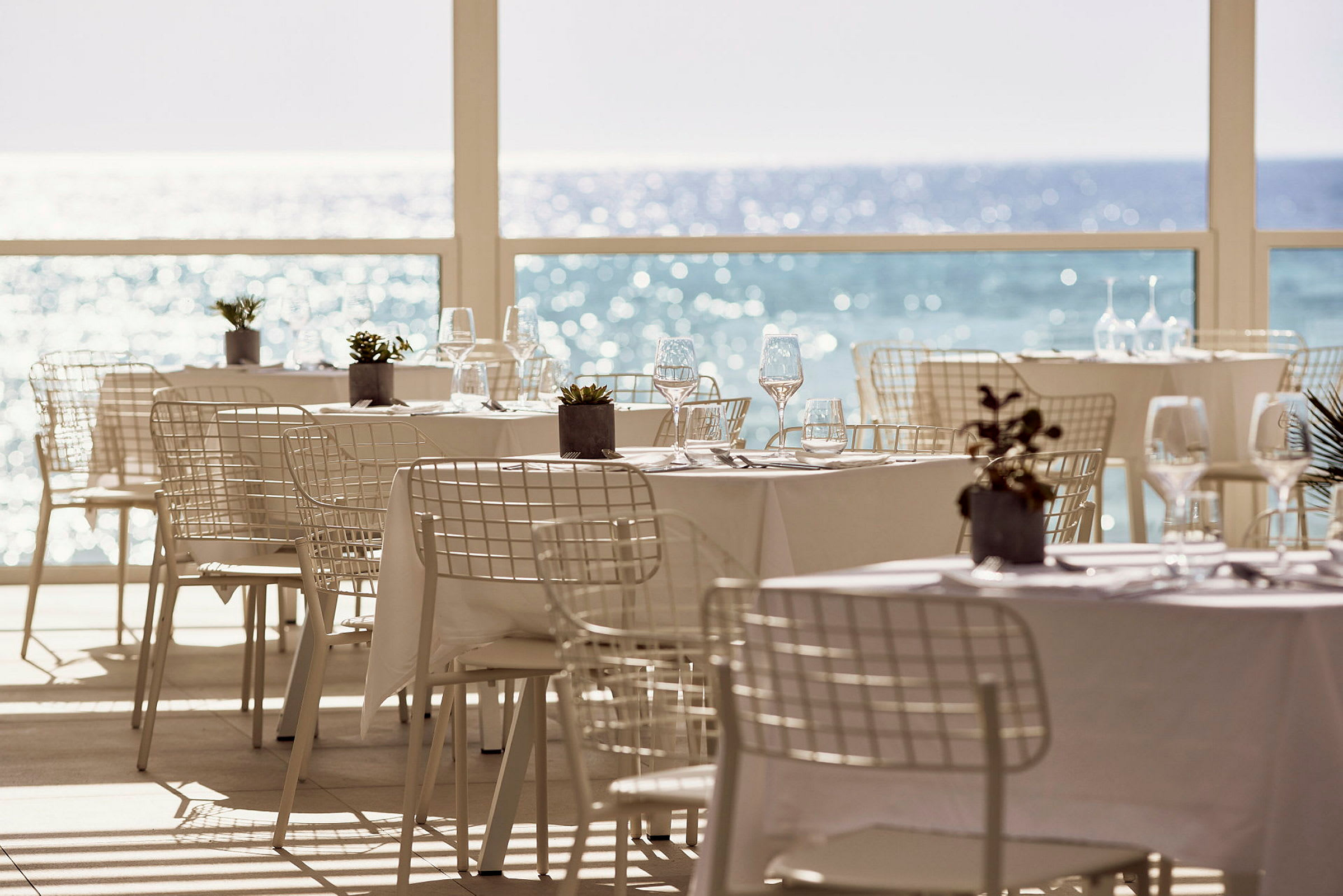 Restaurantborde med hvide duge og stole tæt på et stort vindue med udsigt over havet. (AI-genereret tekst)