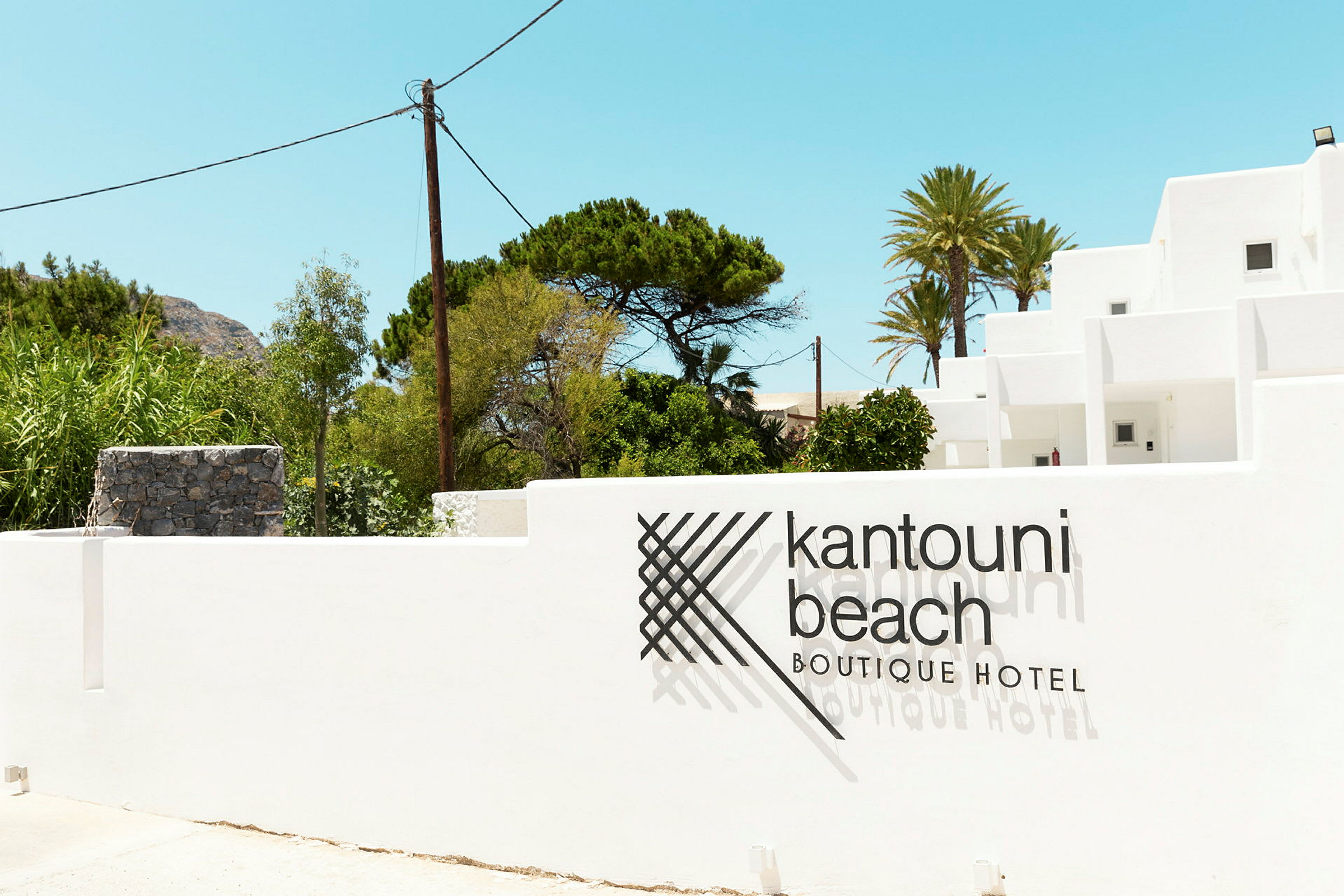 Billede af en hvid bygning med teksten 'Kantouni Beach Boutique Hotel' på muren foran. Grønt og klar himmel i baggrunden. (AI-genereret tekst)