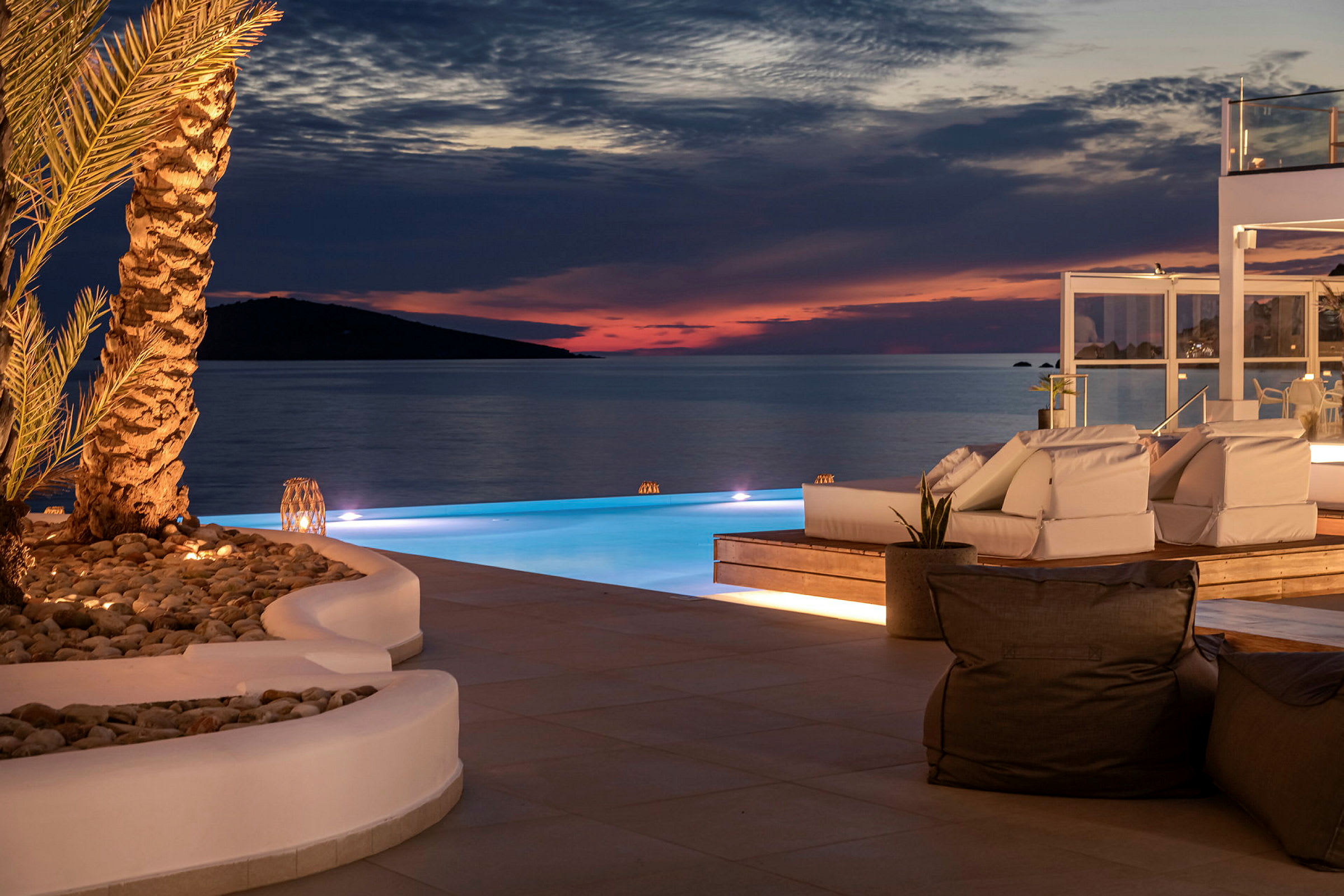Solnedgang ved infinity-pool med loungemøbler og stilfuld belysning. (AI-genereret tekst)