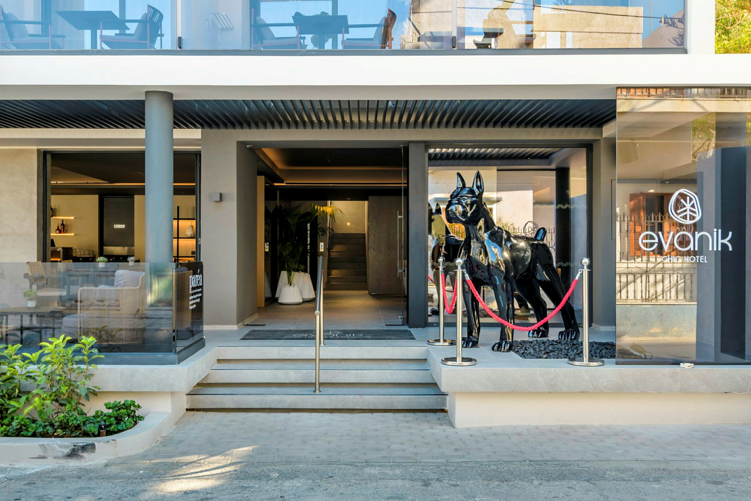 Indgangen til Evanik Chic Hotel med en stor sort skulptur af en hund foran glasdørene. (AI-genereret tekst)