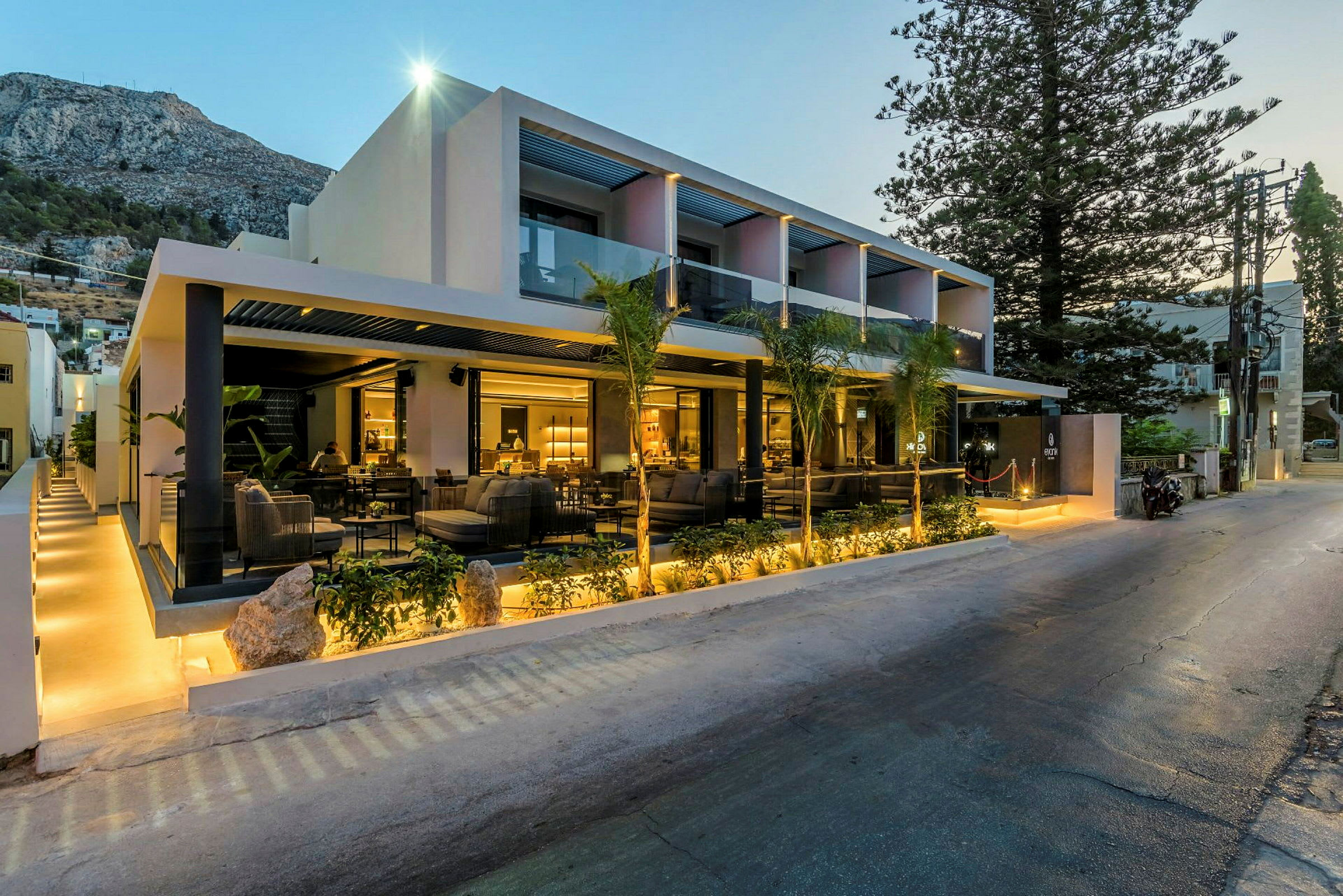 Bygningen er et moderne hotel med et oplyst udendørsområde og omgivende grønt. (AI-genereret tekst)