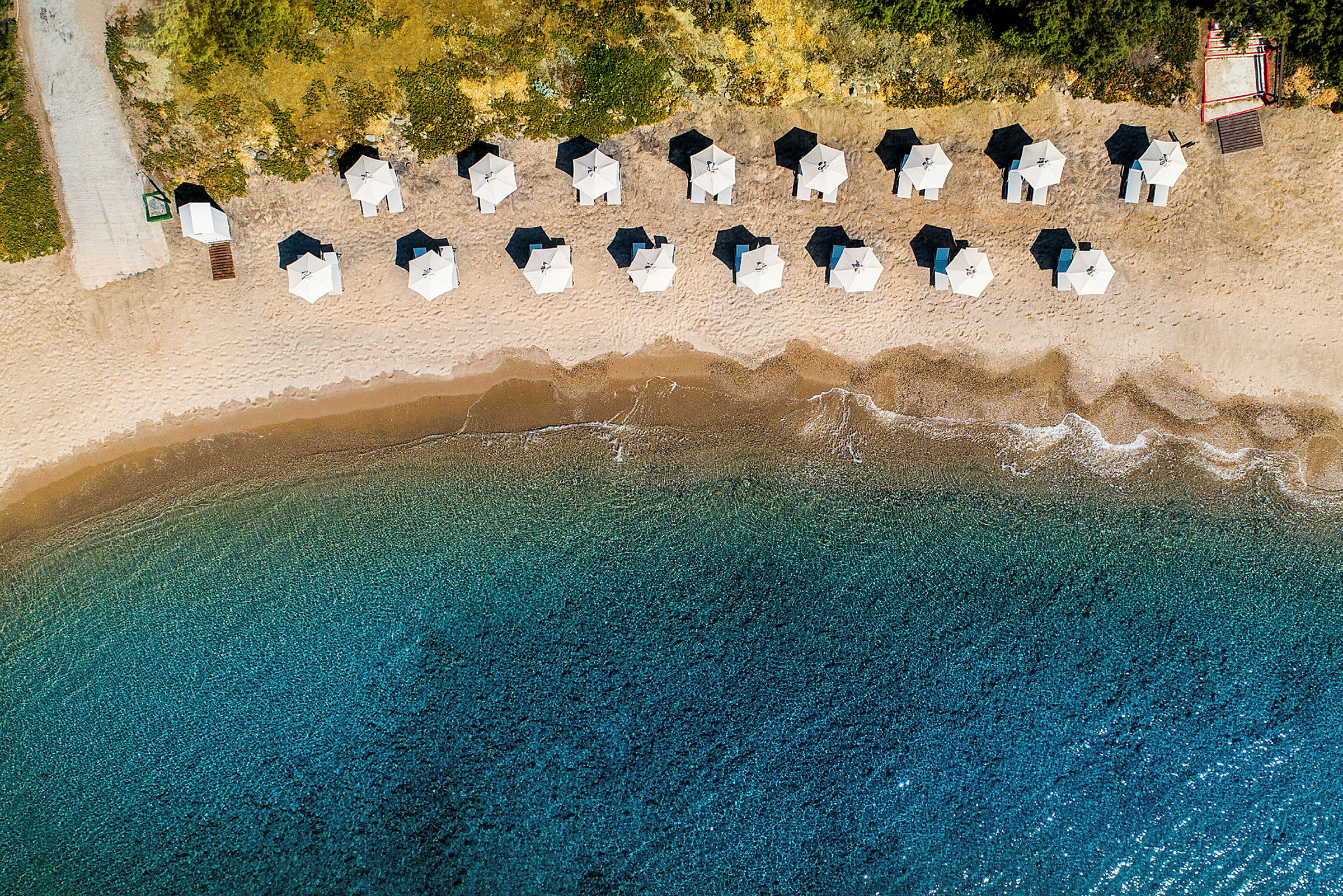 Luftfoto af en sandstrand med parasoller i lige rækker og klart blåt vand. (AI-genereret tekst)
