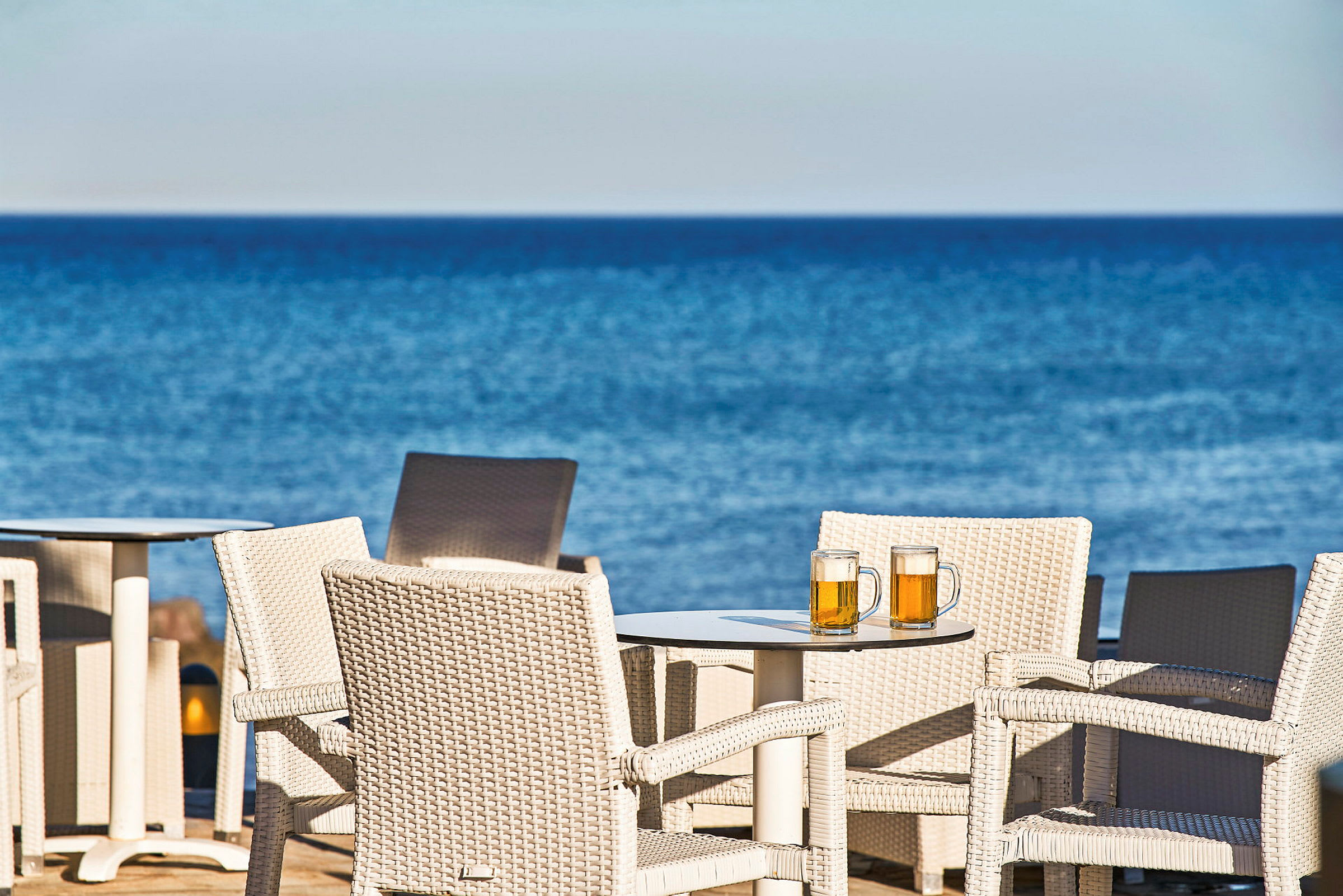 Kurvestole ved et bord med to ølglas, med havet i baggrunden. (AI-genereret tekst)