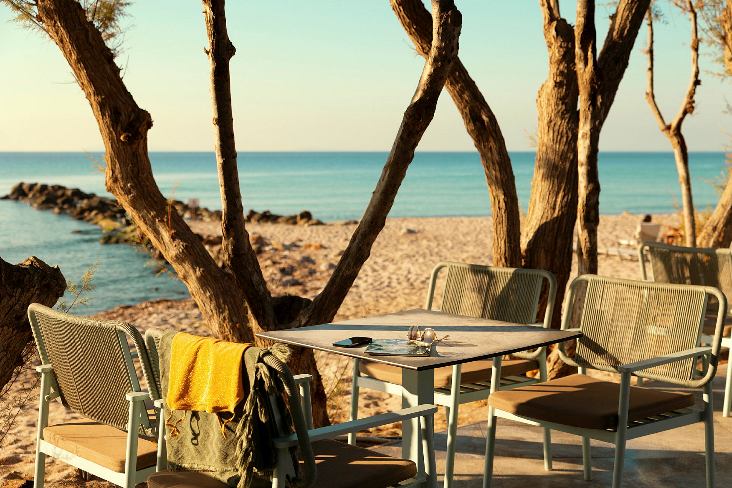 En strandscene med et bord og stole på en terrasse blandt træer, samt udsigt over havet og sandstranden. (AI-genereret tekst)