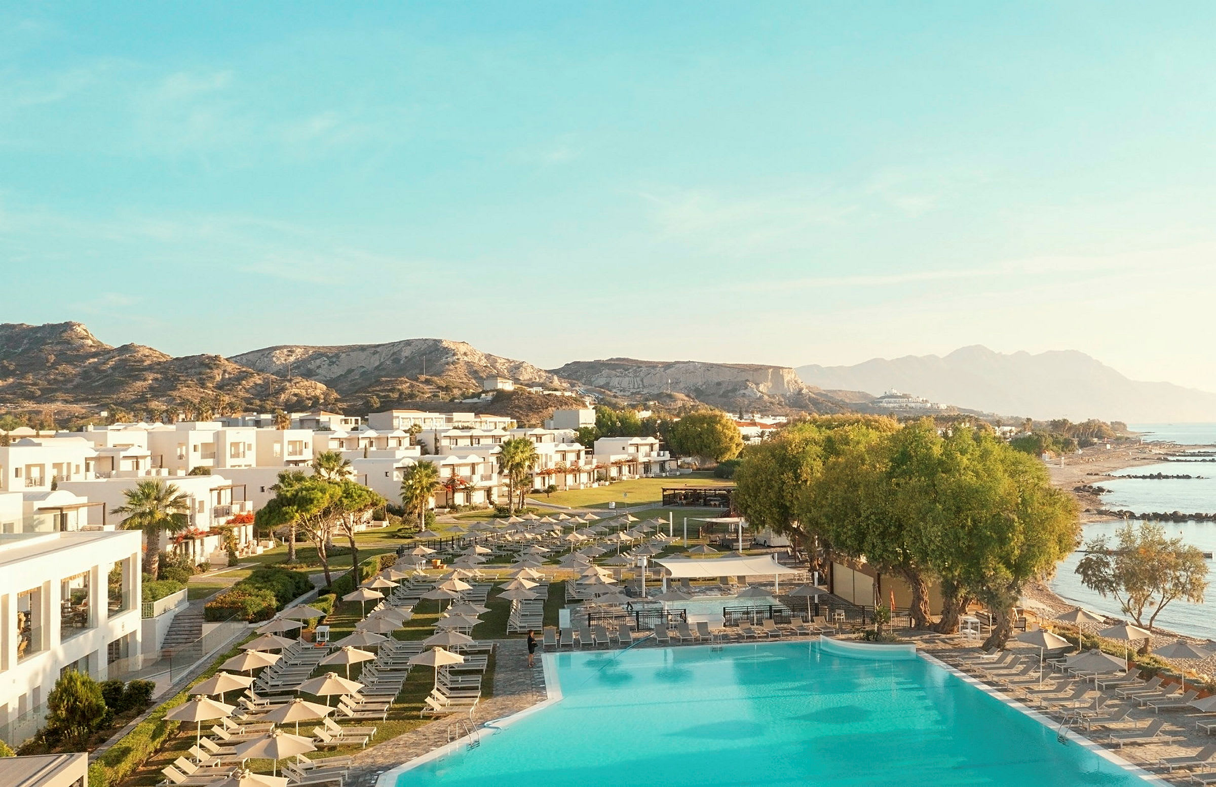 Udsigt over et hotelområde med pool, grønne områder og sandstrande, omgivet af bjerge og hav. (AI-genereret tekst)