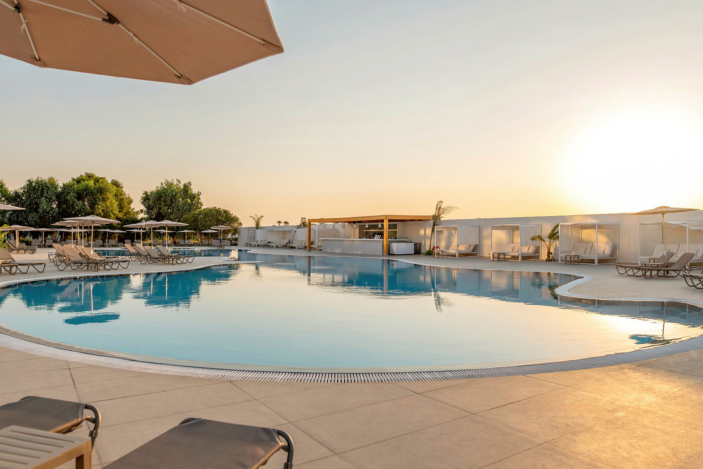 En pool med liggestole og parasoller, omgivet af træer og bygninger ved solnedgang. (AI-genereret tekst)
