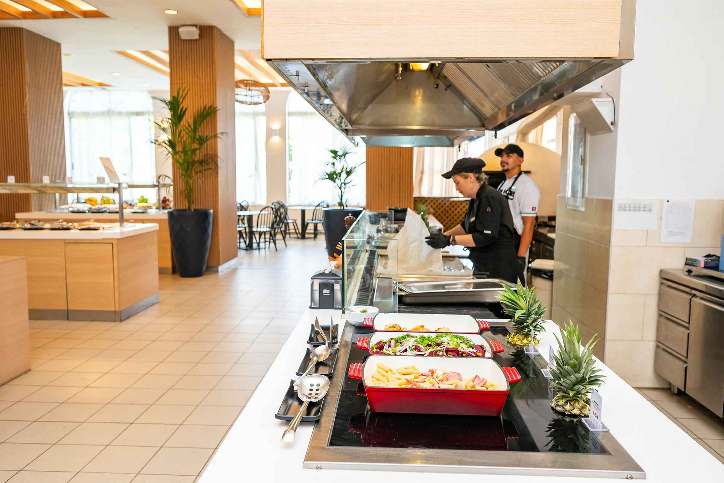 Buffetområde med mad anrettet i farverige serveringsfade og køkkenpersonale ved arbejdsstation. (AI-genereret tekst)