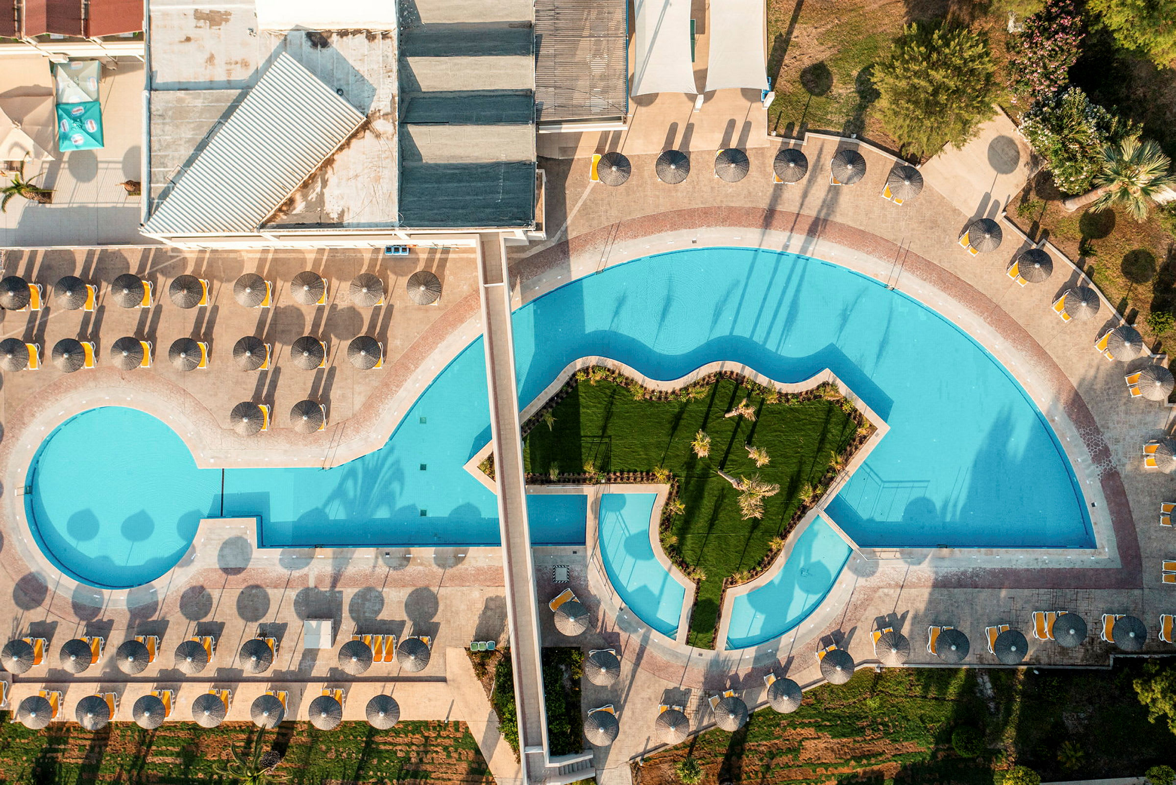 Luftfoto af en swimmingpool med en græsklædt ø i midten, liggestole og parasoller omkring. (AI-genereret tekst)