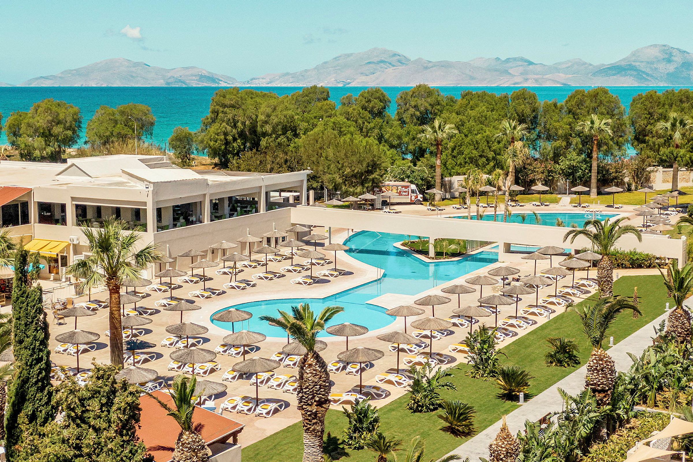 Hotelområde med pool, liggestole og parasoller, omgivet af grønne områder med havet i baggrunden. (AI-genereret tekst)