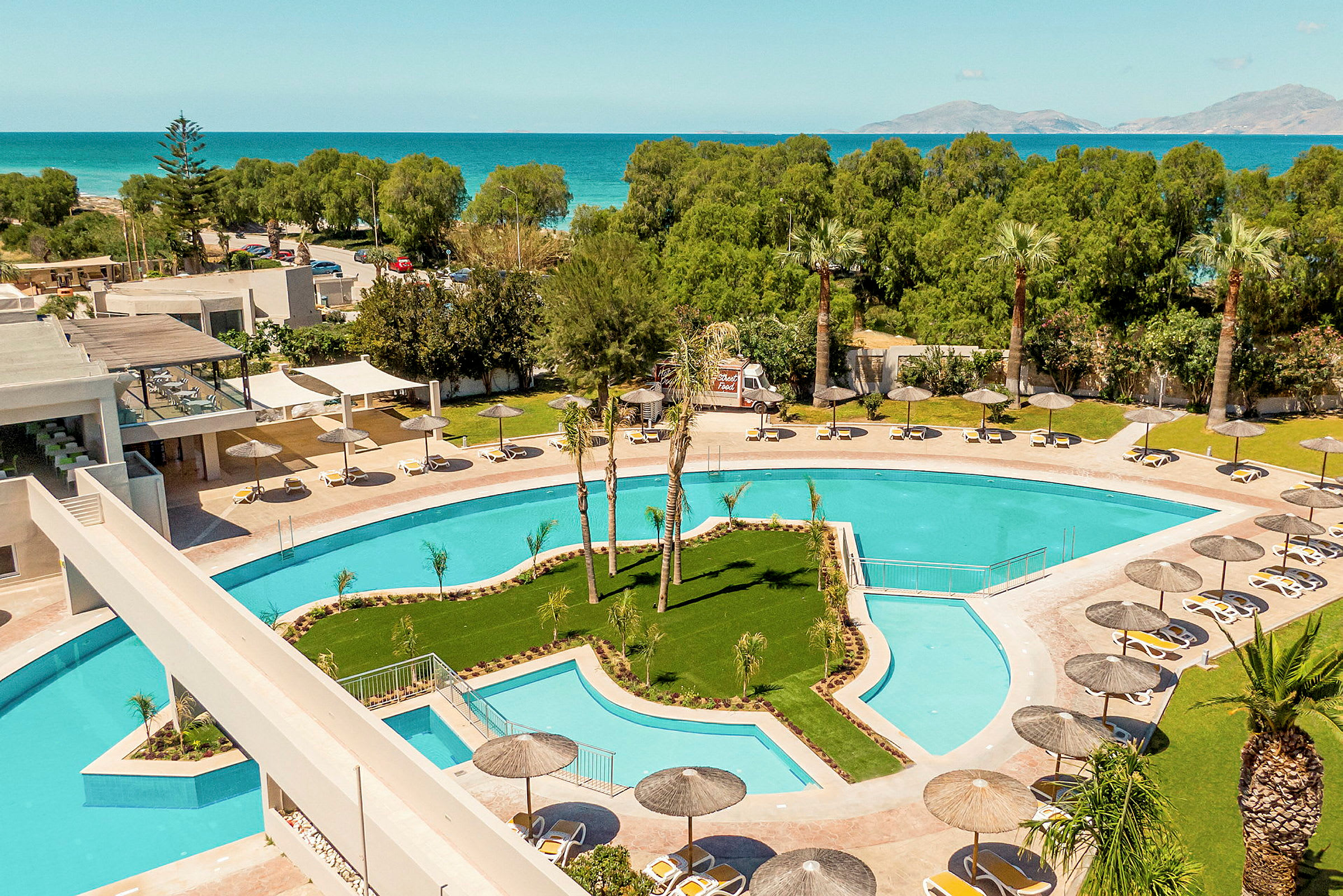 En hotelpool med liggestole og parasoller, omgivet af træer og med udsigt til havet. (AI-genereret tekst)