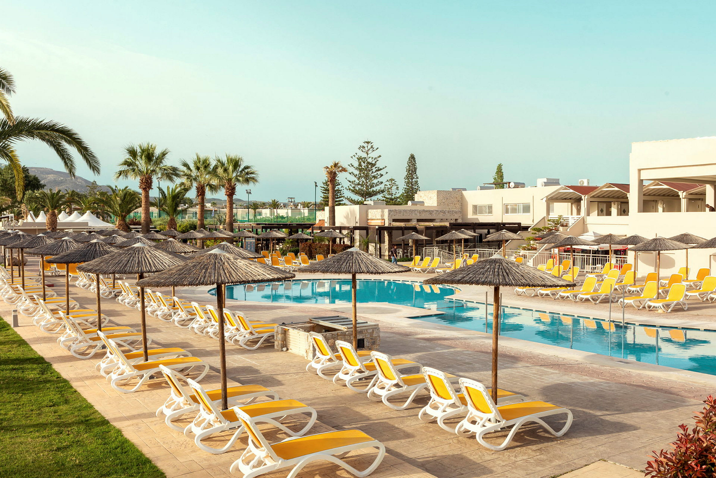 Liggestole og parasoller omkring hotellets swimmingpool med palmer og bygninger i baggrunden. (AI-genereret tekst)