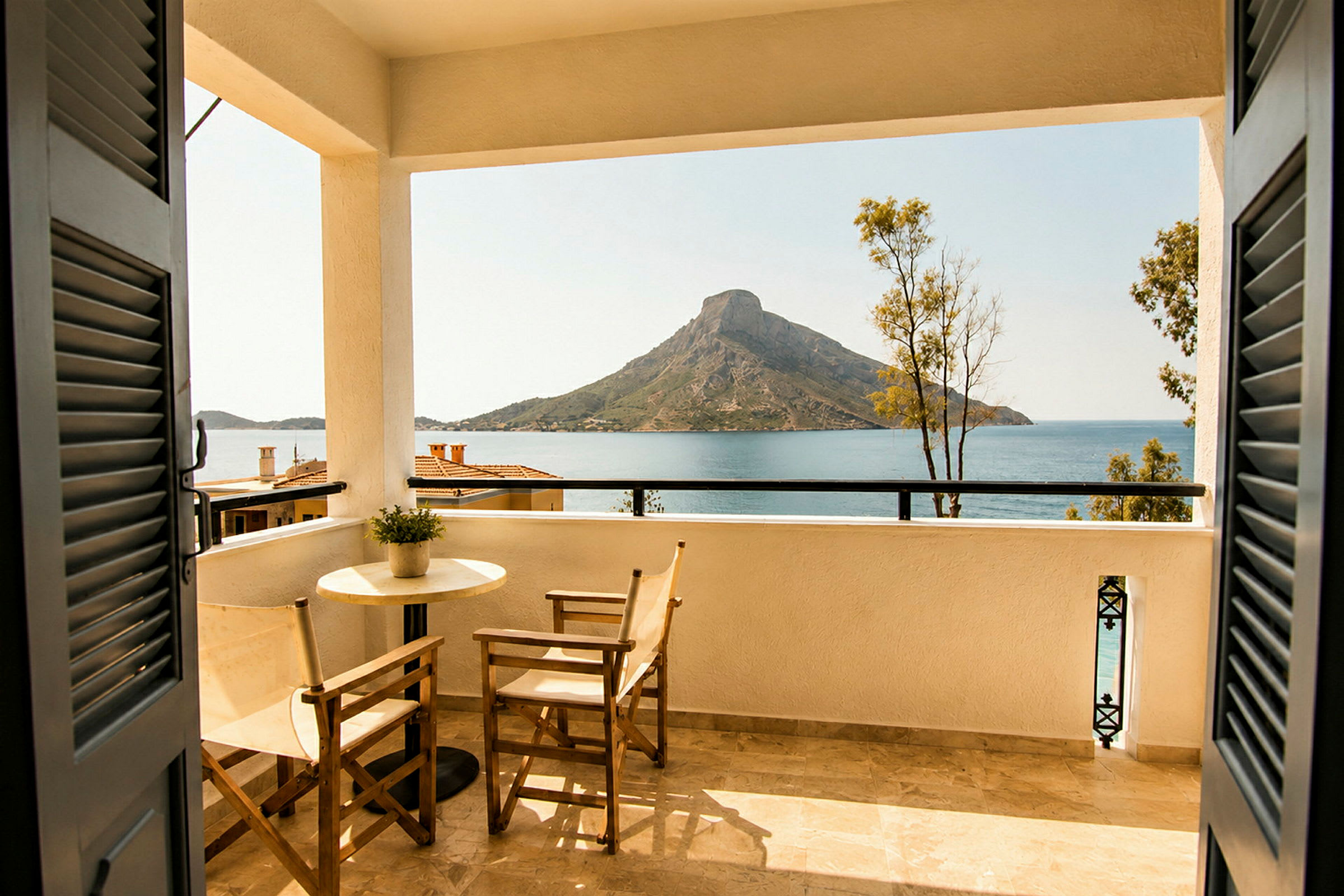 Udsigt fra en balkon med bord og stole mod havet og en bjergrig ø. (AI-genereret tekst)