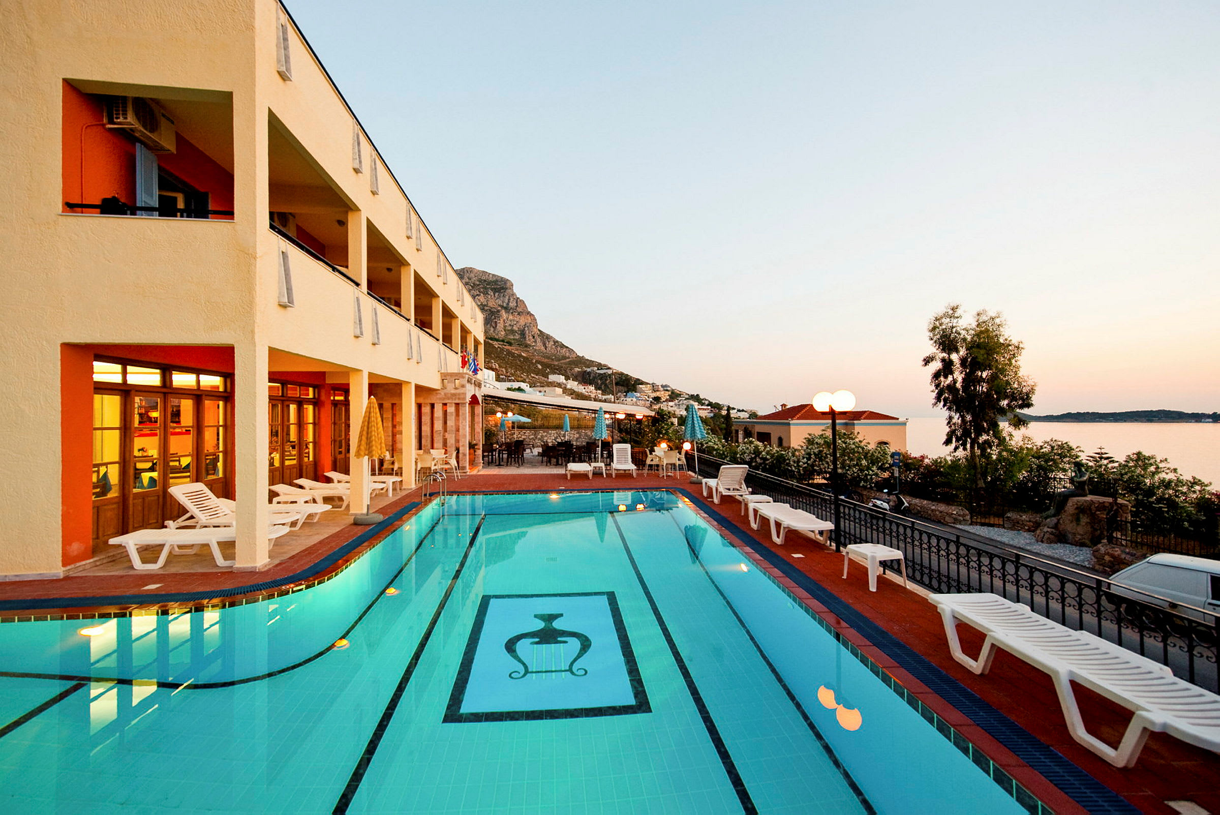 Udendørs swimmingpool med liggestole, omgivet af et hotel og vegetation ved solnedgang. (AI-genereret tekst)