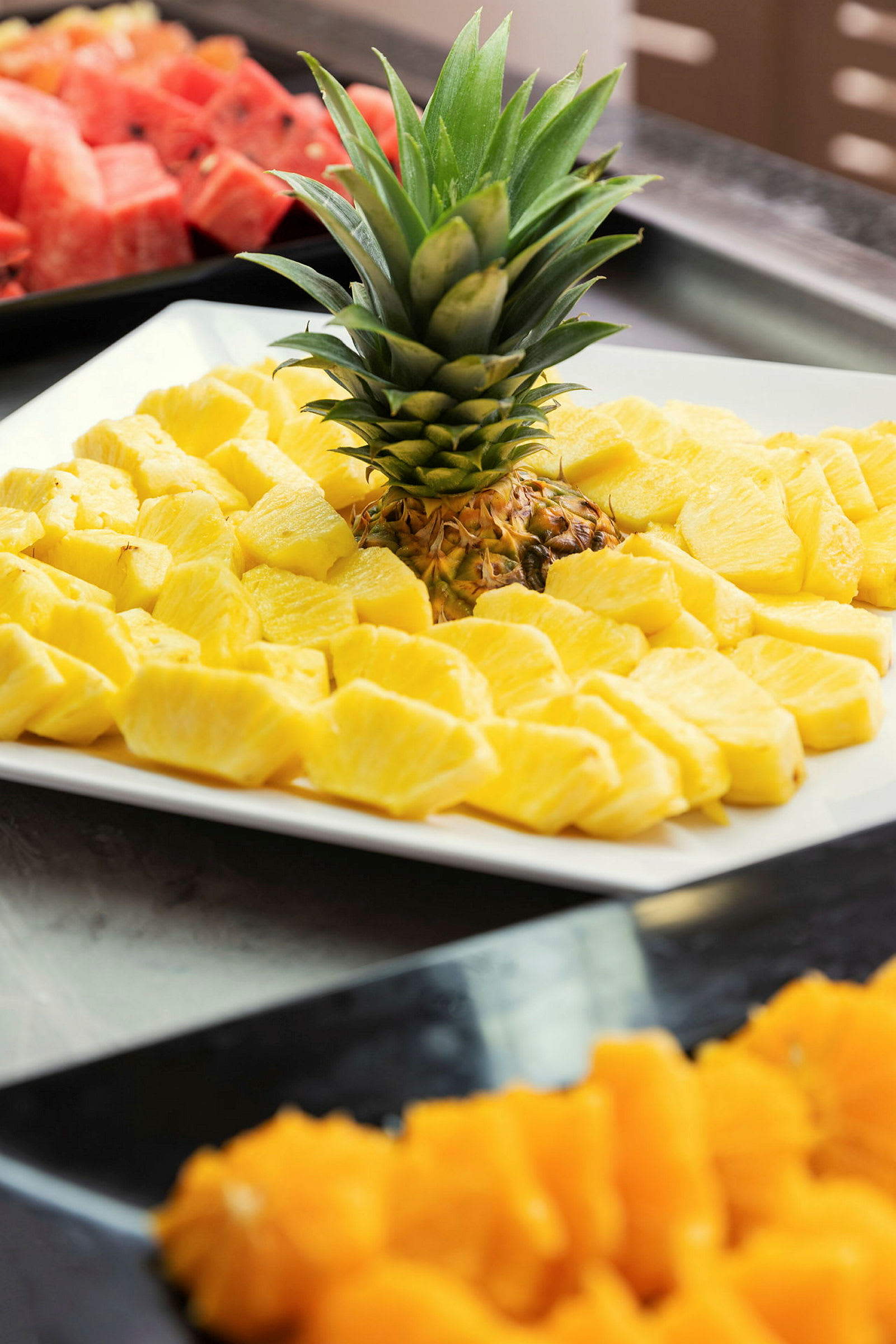 Et nærbillede af en tallerken med skiver af frisk ananas og dens grønne krone, med andre frugter i baggrunden. (AI-genereret tekst)