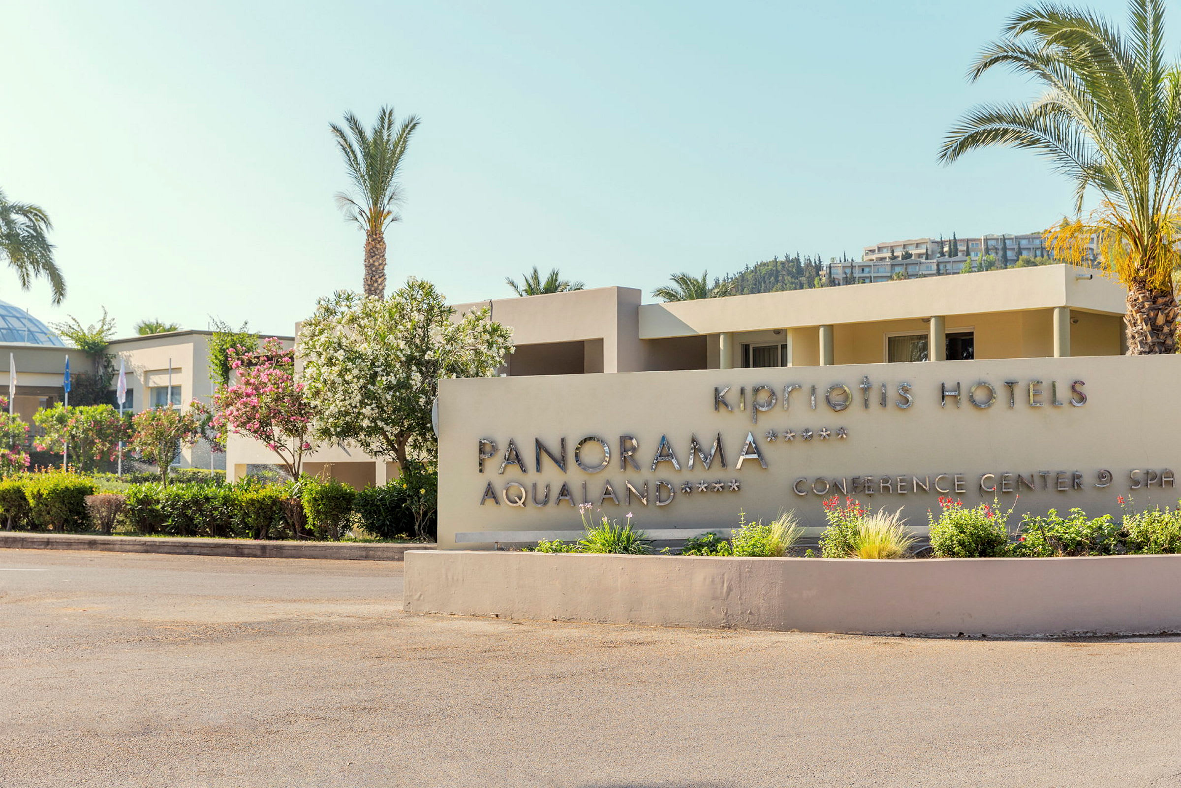 Hotellets skilt med teksten 'Kipriotis Hotels Panorama Aqualand Conference Center & Spa', omgivet af planter og palmer. (AI-genereret tekst)