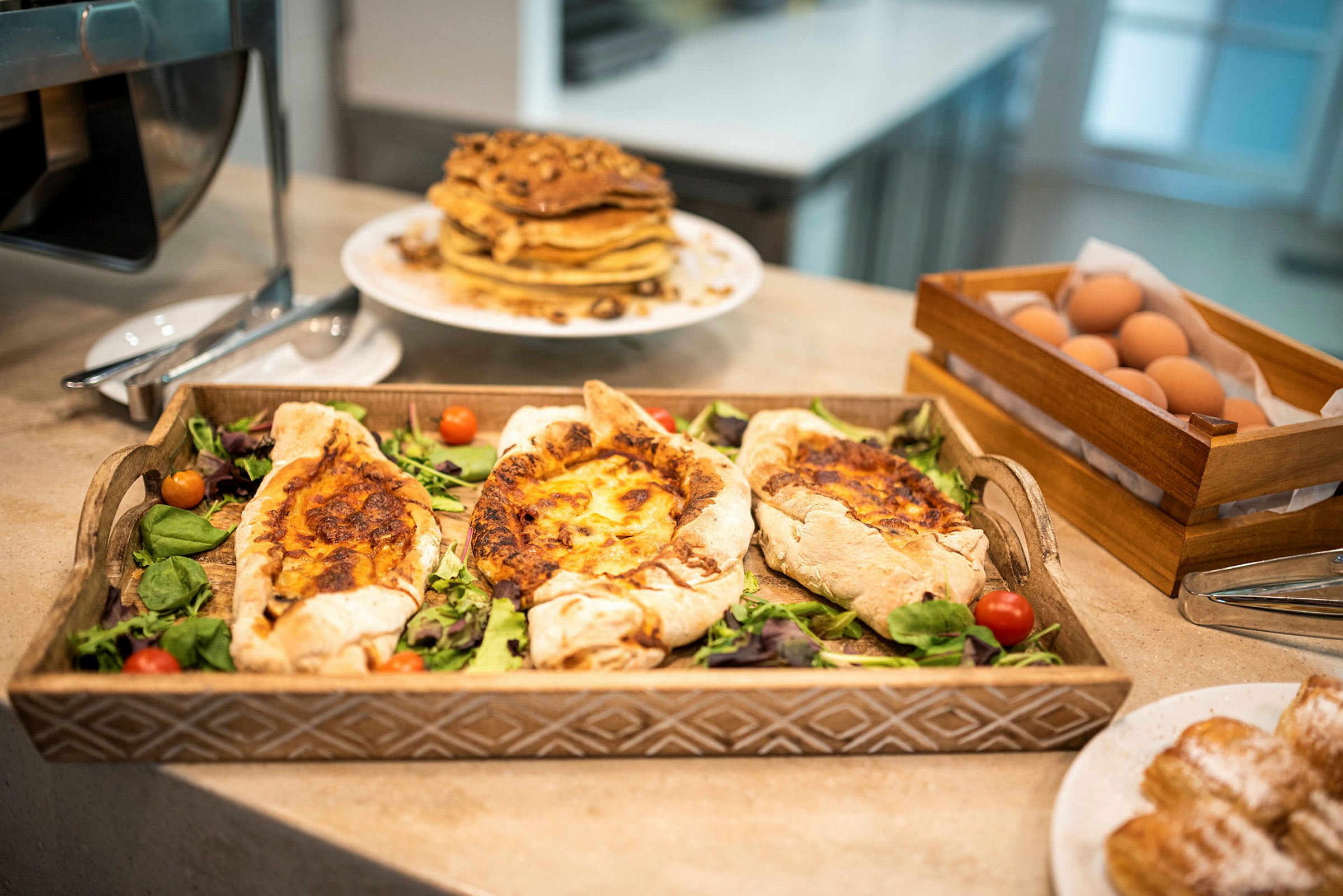 Et fad med fyldte brød på salatblade, omgivet af en tallerken med pandekager, nybagte croissanter og en trækasse med æg. (AI-genereret tekst)