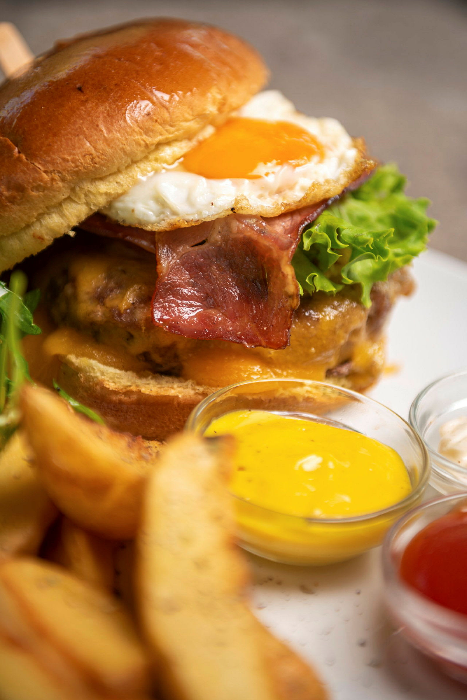 En burger med bacon, ost, salat og spejlæg serveres med kartoffelbåde og forskellige saucer. (AI-genereret tekst)
