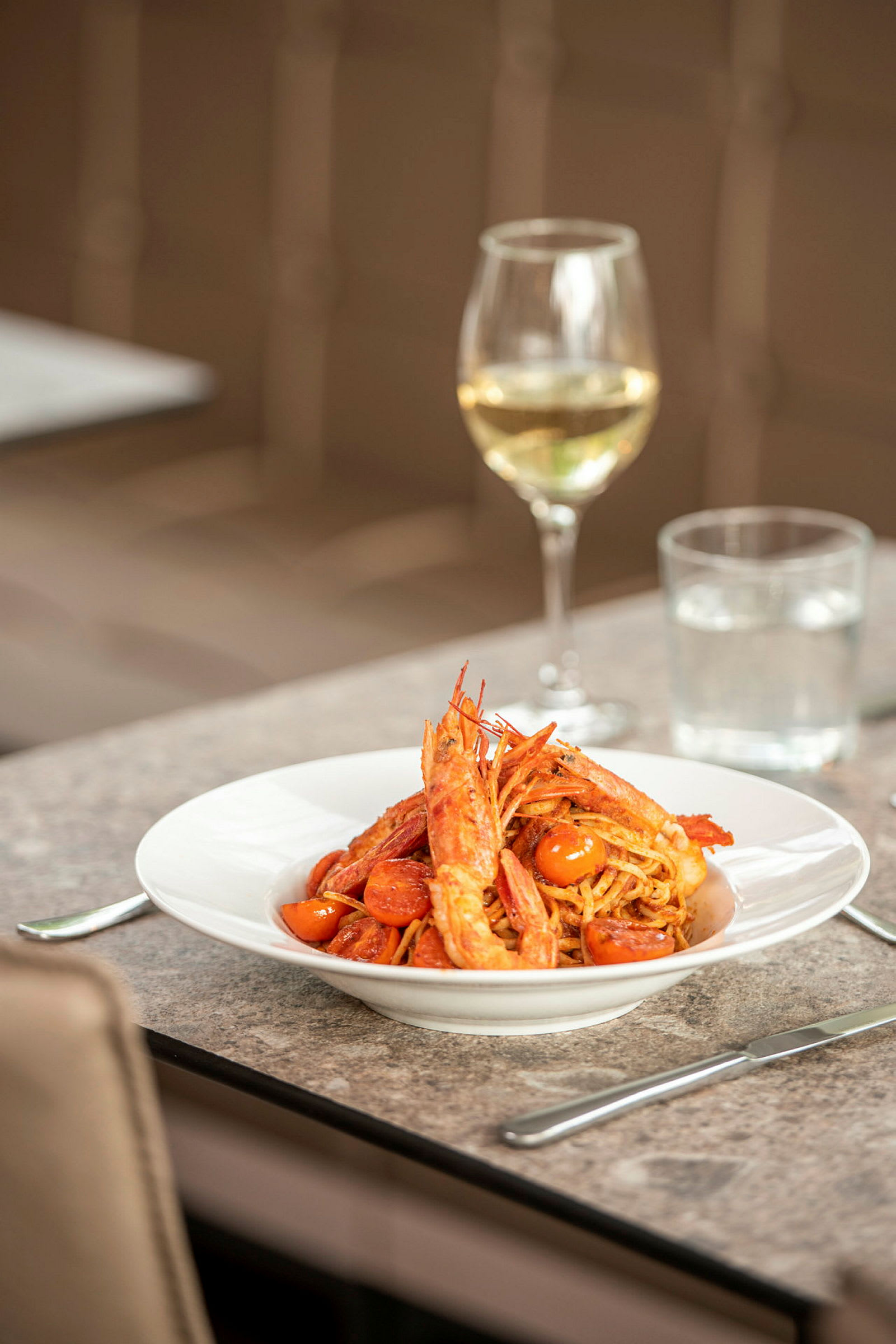 En tallerken med pasta, rejer og tomater står på et bord. I baggrunden ses et glas hvidvin. (AI-genereret tekst)