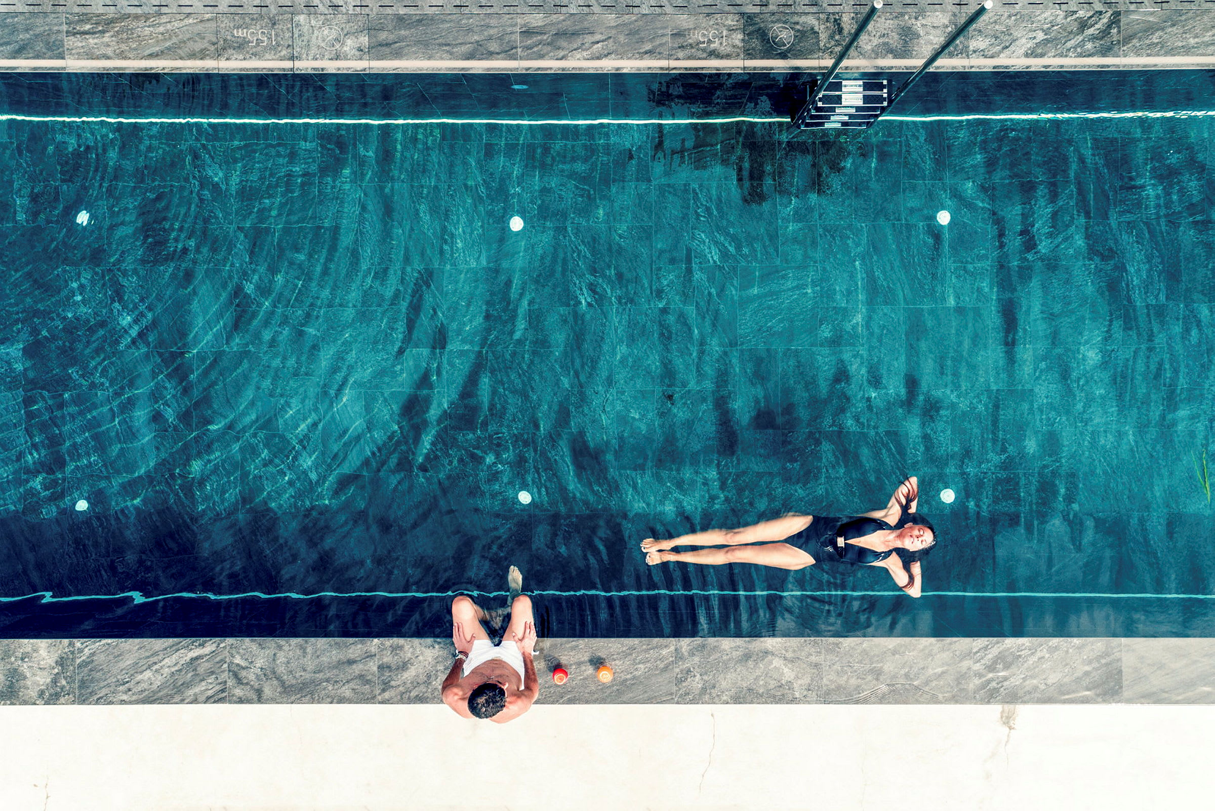 Person bader i en pool ved siden af en, der sidder på kanten. (AI-genereret tekst)