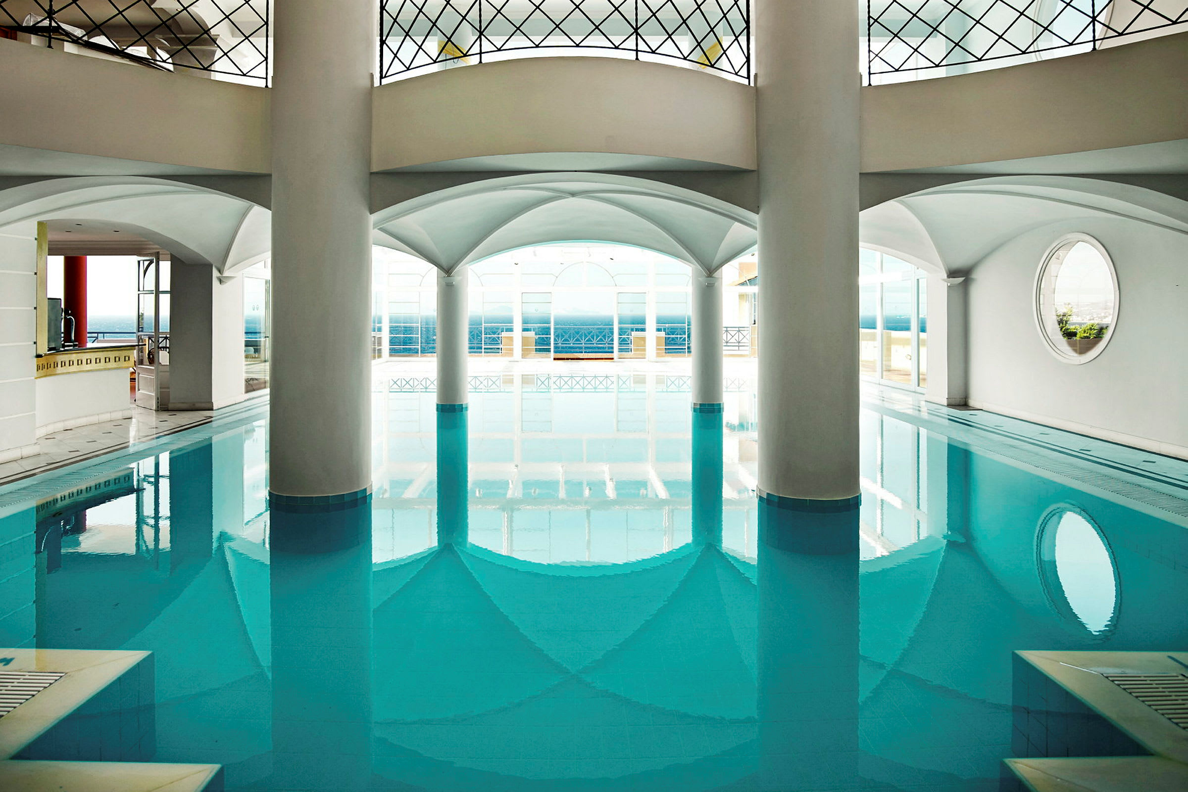 En indendørs pool omgivet af hvide søjler og buede lofter, med udsigt til en terrasse og havet i baggrunden. (AI-genereret tekst)