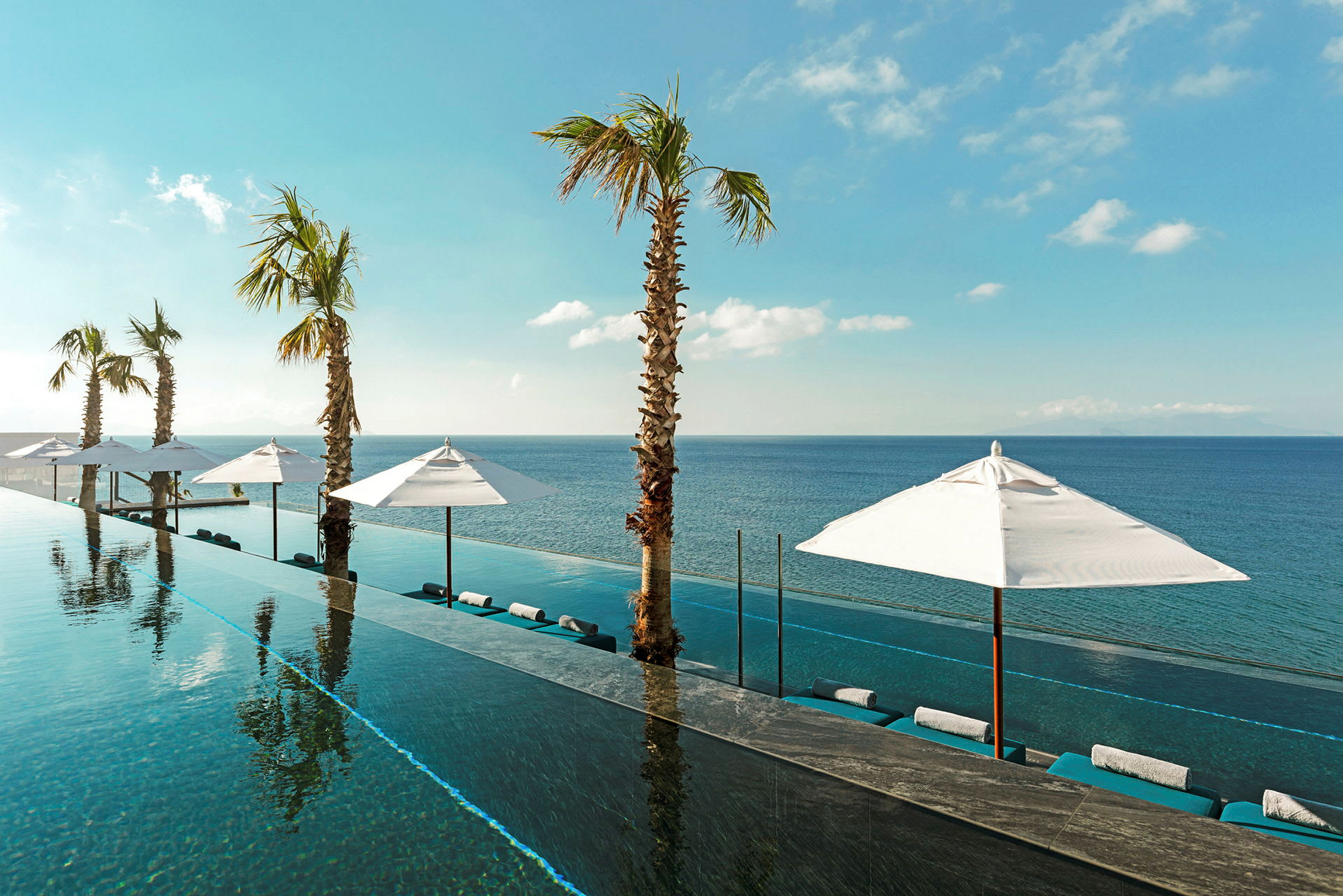 En infinitypool med udsigt over havet, omgivet af parasoller og palmer under en klar blå himmel. (AI-genereret tekst)