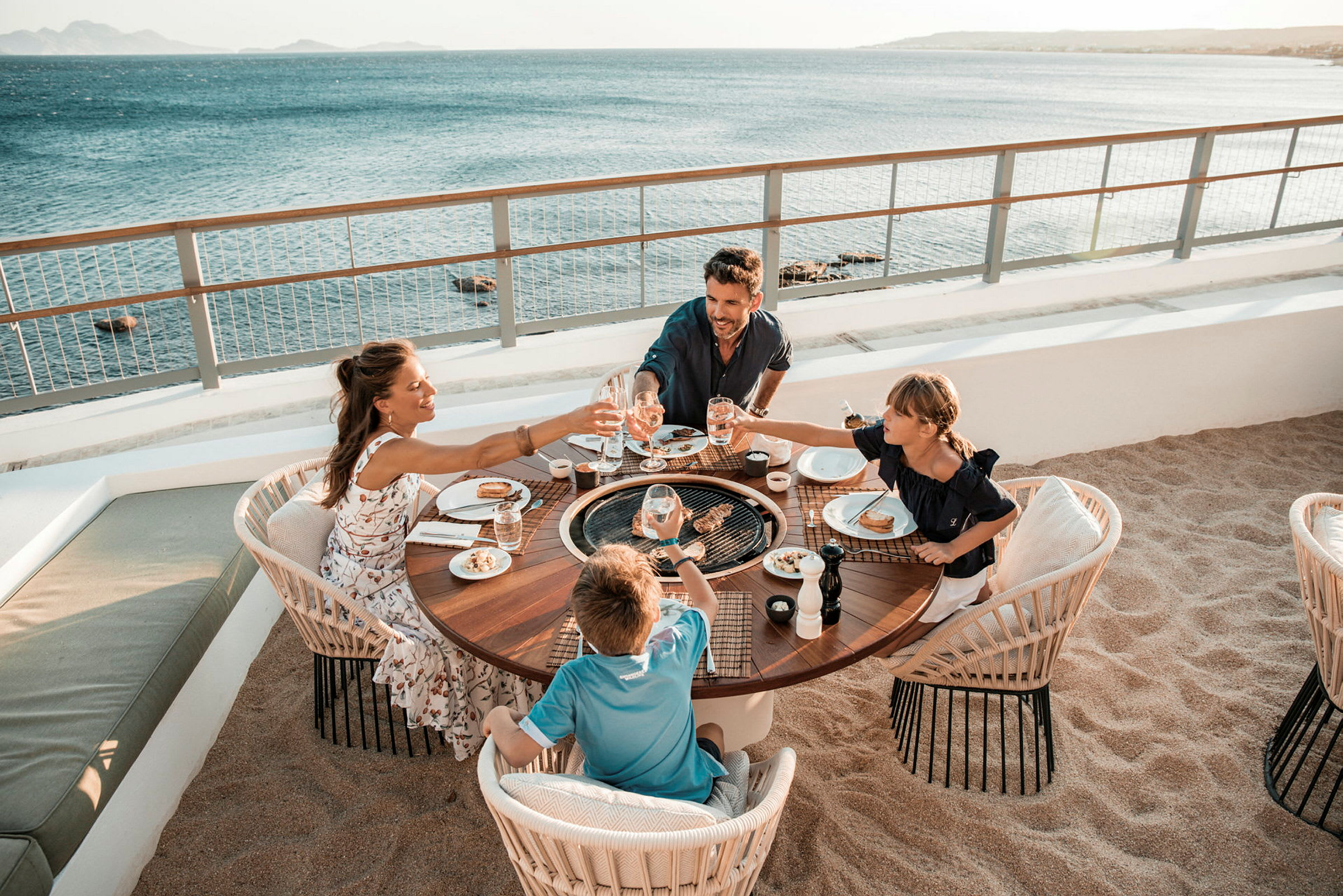En familie sidder ved et rundt bord udendørs ved stranden og nyder et måltid med udsigt over havet. (AI-genereret tekst)