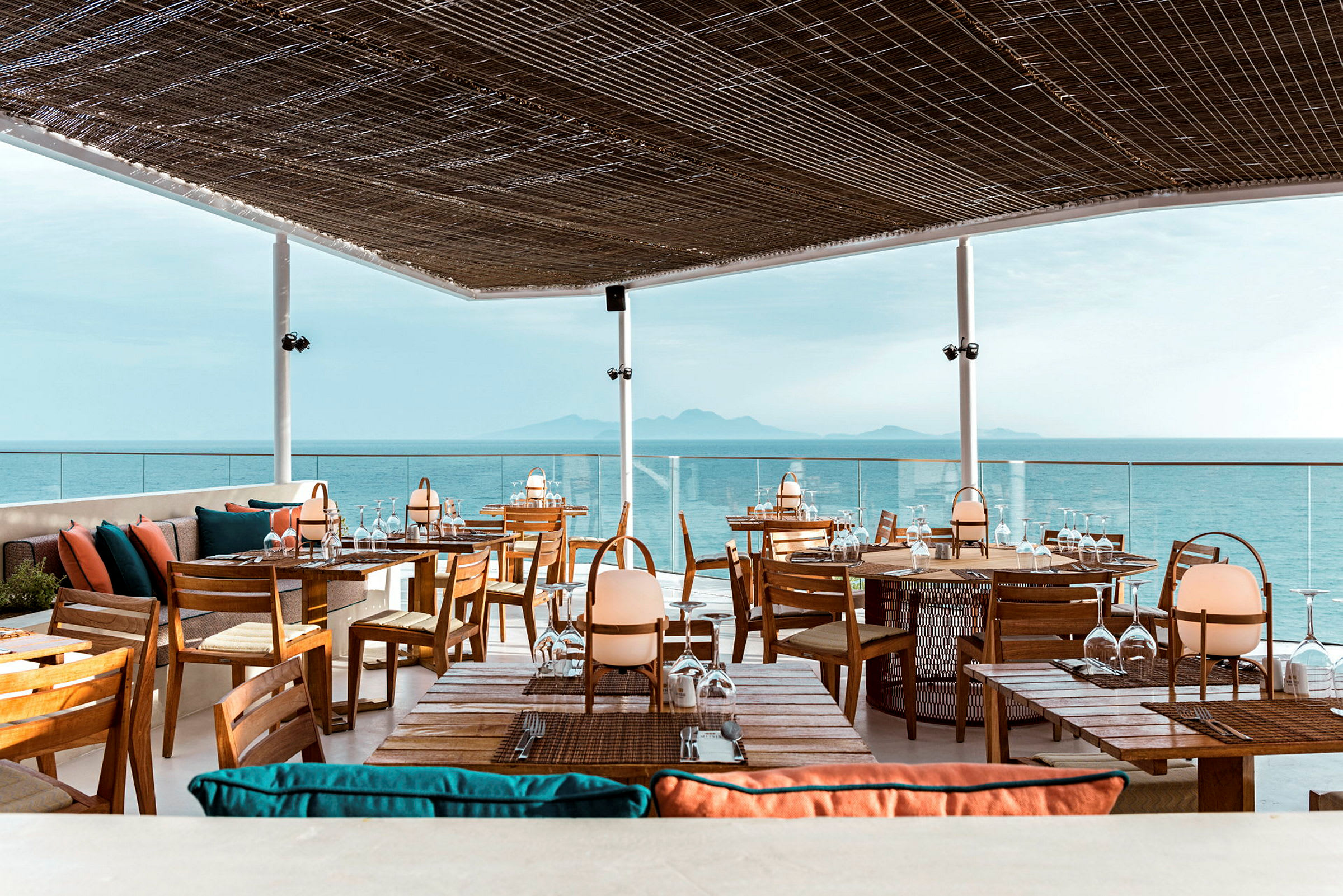 En restaurant med tagterrasse med udsigt over havet. Møblerne er lavet af træ med farverige puder, og taget består af et flettet materiale, der lader lys komme igennem. (AI-genereret tekst)