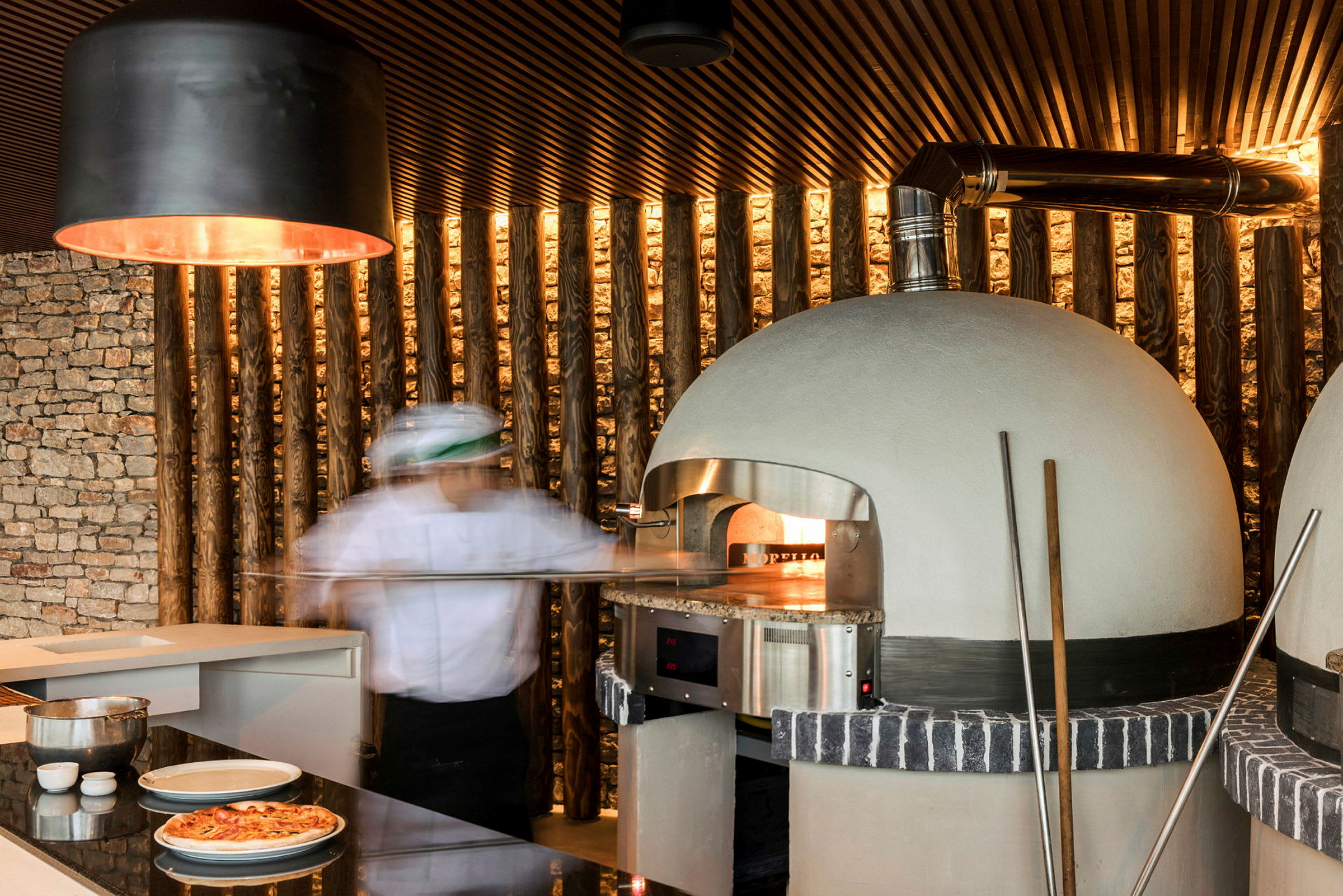 En pizzabager foran en brændefyret ovn med et moderne køkken i baggrunden. (AI-genereret tekst)