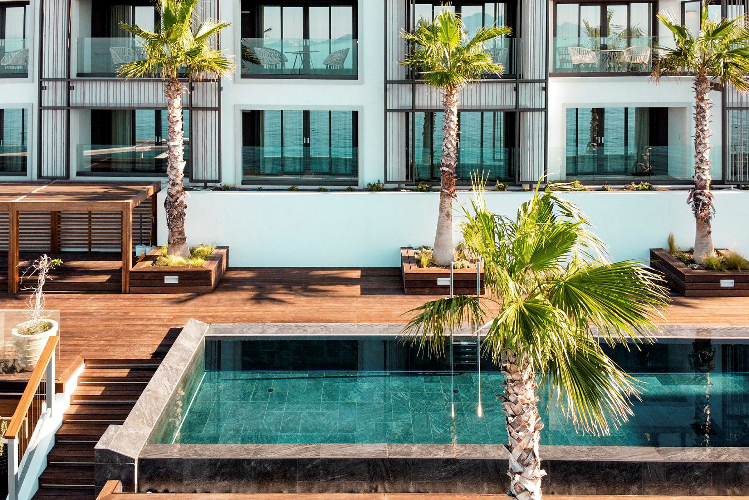 En pool omgivet af træterrasser og palmer foran et moderne hotel. (AI-genereret tekst)