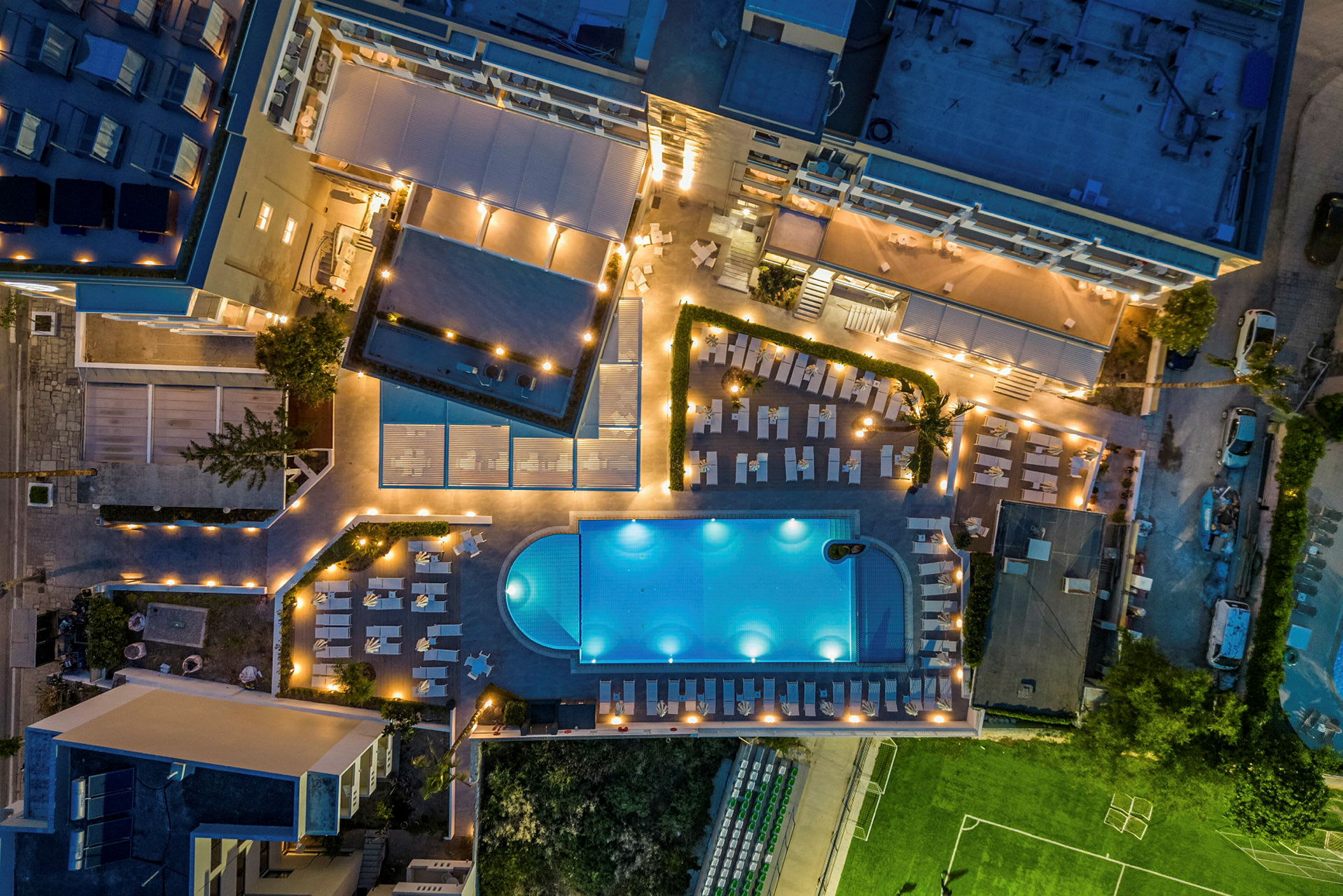 Luftfoto af hotelområde med pool, liggestole og grønne områder, oplyst om aftenen. (AI-genereret tekst)