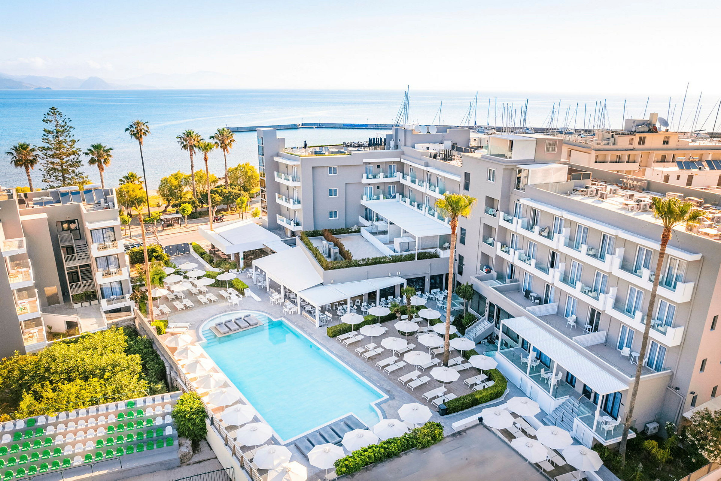 Et hotelområde med pool, parasoller og liggestole omgivet af bygninger. Hotellet ligger nær havet med en strand og en lystbådehavn i baggrunden. (AI-genereret tekst)