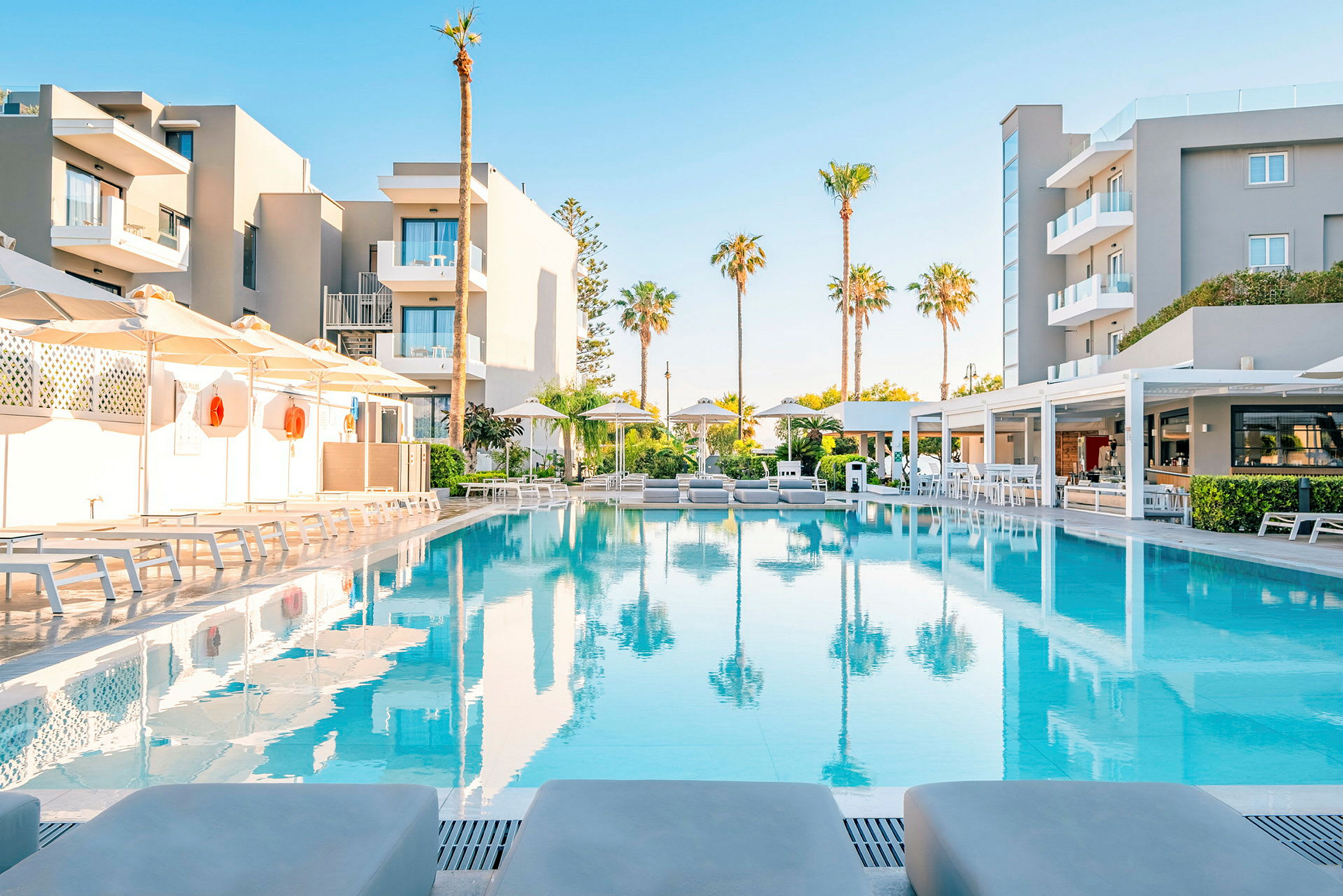 En moderne hotelbygning med en oplyst pool foran, flankeret af solsenge og parasoller. (AI-genereret tekst)