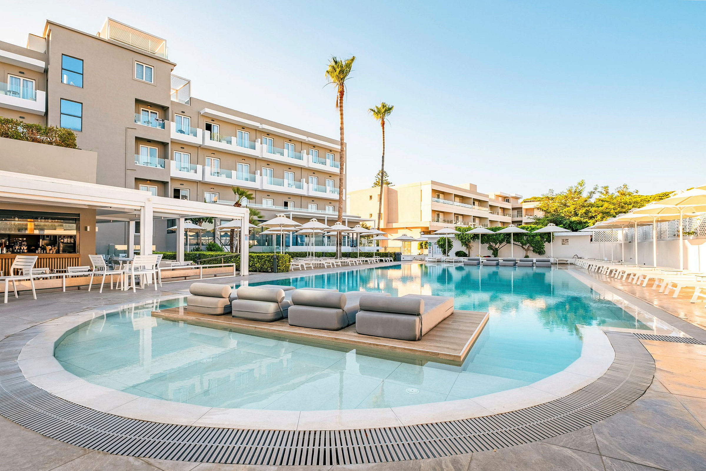 En hotelpool omgivet af liggestole og parasoller, med en fleretagers bygning i baggrunden. (AI-genereret tekst)