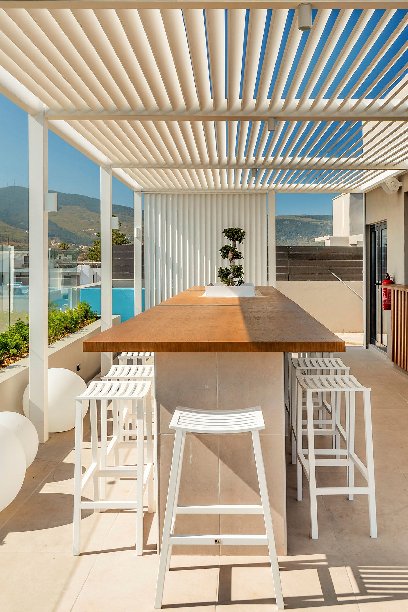 Udendørs serveringsområde med et stort træbord omgivet af hvide barstole og en pergola til skygge. Bjerglandskab kan anes i baggrunden. (AI-genereret tekst)